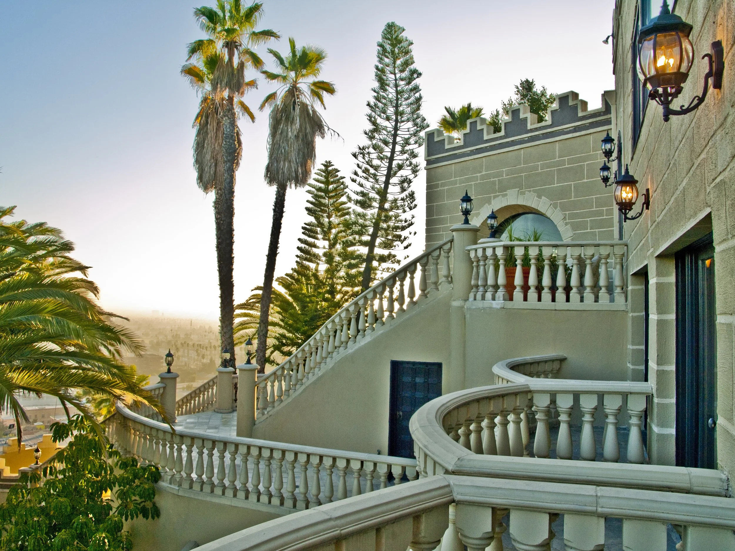 Presented by Missig Team, a real estate team at Berkshire Hathaway HomeServices in Beverly Hills and Los Angeles.  Exterior view of a building with a curved staircase, balustrades, potted plants, and decorative lamps, surrounded by tall palm trees an