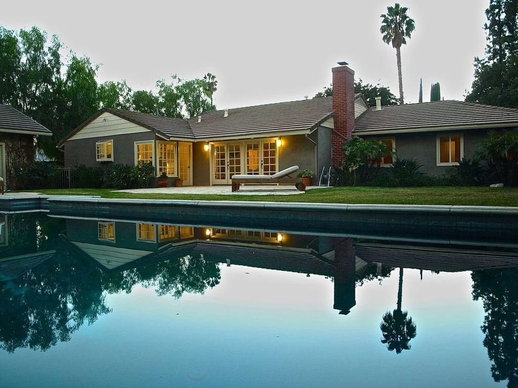 Presented by Missig Team, a real estate team at Berkshire Hathaway HomeServices California Properties in Beverly Hills and Los Angeles. A house with lit windows and outdoor lighting, viewed from a swimming pool, with trees and tall palm trees in the
