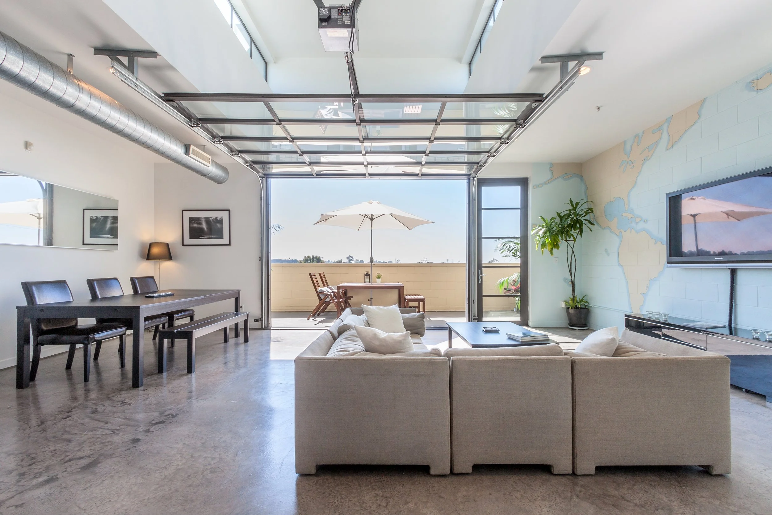 Presented by Missig Team, a real estate team at Berkshire Hathaway HomeServices California Properties in Beverly Hills and Los Angeles. Living room with large glass garage door leading to balcony with outdoor dining table and umbrella, beige sofa, wa