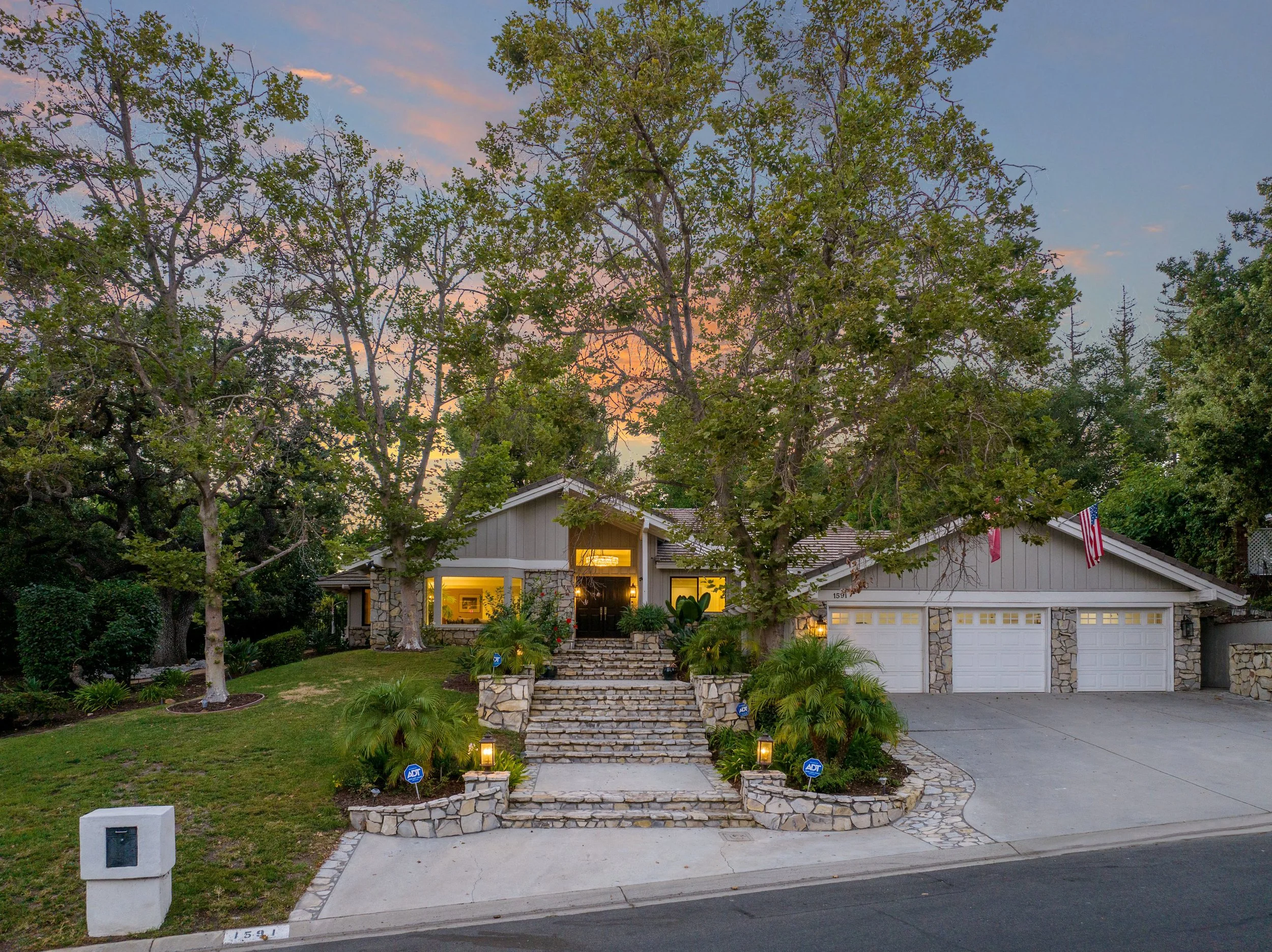 Presented by Missig Team, a real estate team at Berkshire Hathaway HomeServices California Properties in Beverly Hills and Los Angeles. Front view of a house with a stone staircase leading to the front door, surrounded by trees and greenery, with Ame