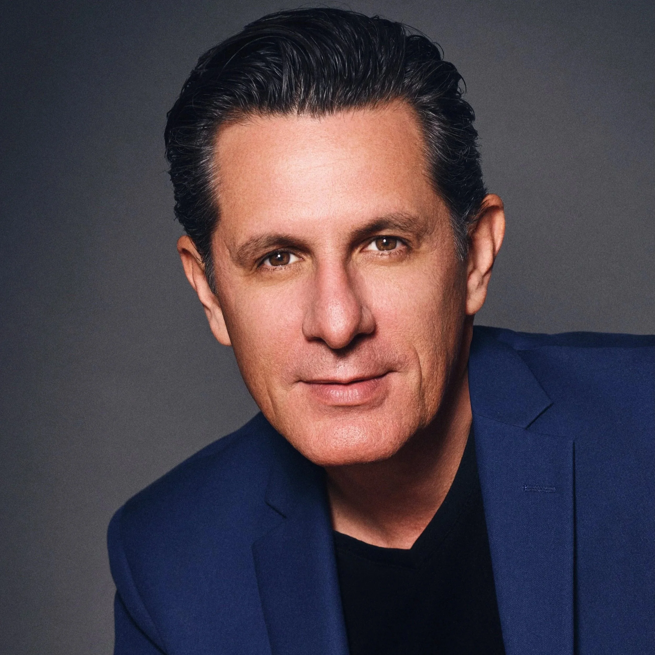 A man realtor with dark brown hair styled back, wearing a dark blue blazer and a black shirt, poses against a dark gray background.