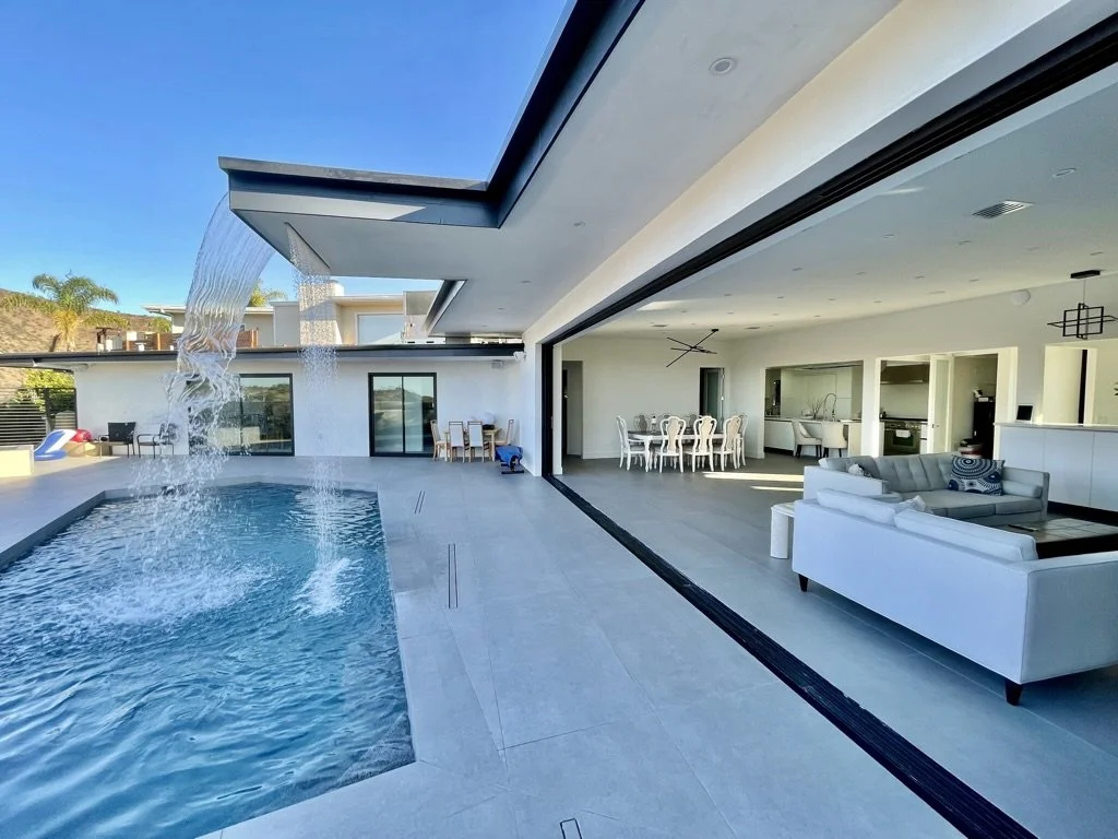 Presented by Missig Team, a real estate team at Berkshire Hathaway HomeServices California Properties in Beverly Hills and Los Angeles. Modern outdoor pool area with waterfall feature, open living space with white sofas and dining table, and a clear