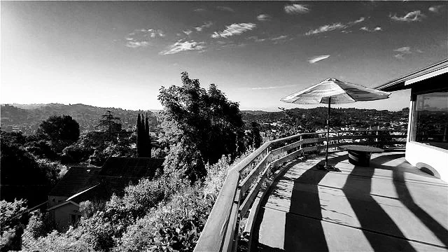 Presented by Missig Team, a real estate team at Berkshire Hathaway HomeServices California Properties in Beverly Hills and Los Angeles. A black and white photo of a curved balcony with a patio umbrella, overlooking a hilly landscape with trees and ho