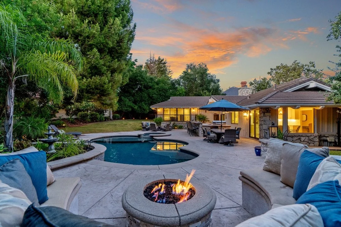 Presented by Missig Team, a real estate team at Berkshire Hathaway HomeServices California Properties in Beverly Hills and Los Angeles. View of a Westlake Village backyard patio at sunset with a pool, fire pit, lounge chairs, outdoor dining area, and