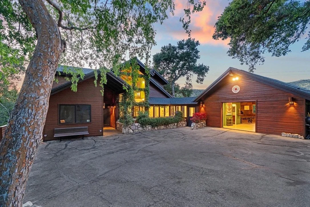 Presented by Missig Team, a real estate team at Berkshire Hathaway HomeServices California Properties in Beverly Hills and Los Angeles. A large house with wooden exterior and a garage door, illuminated from inside, set in a wooded area during sunset.