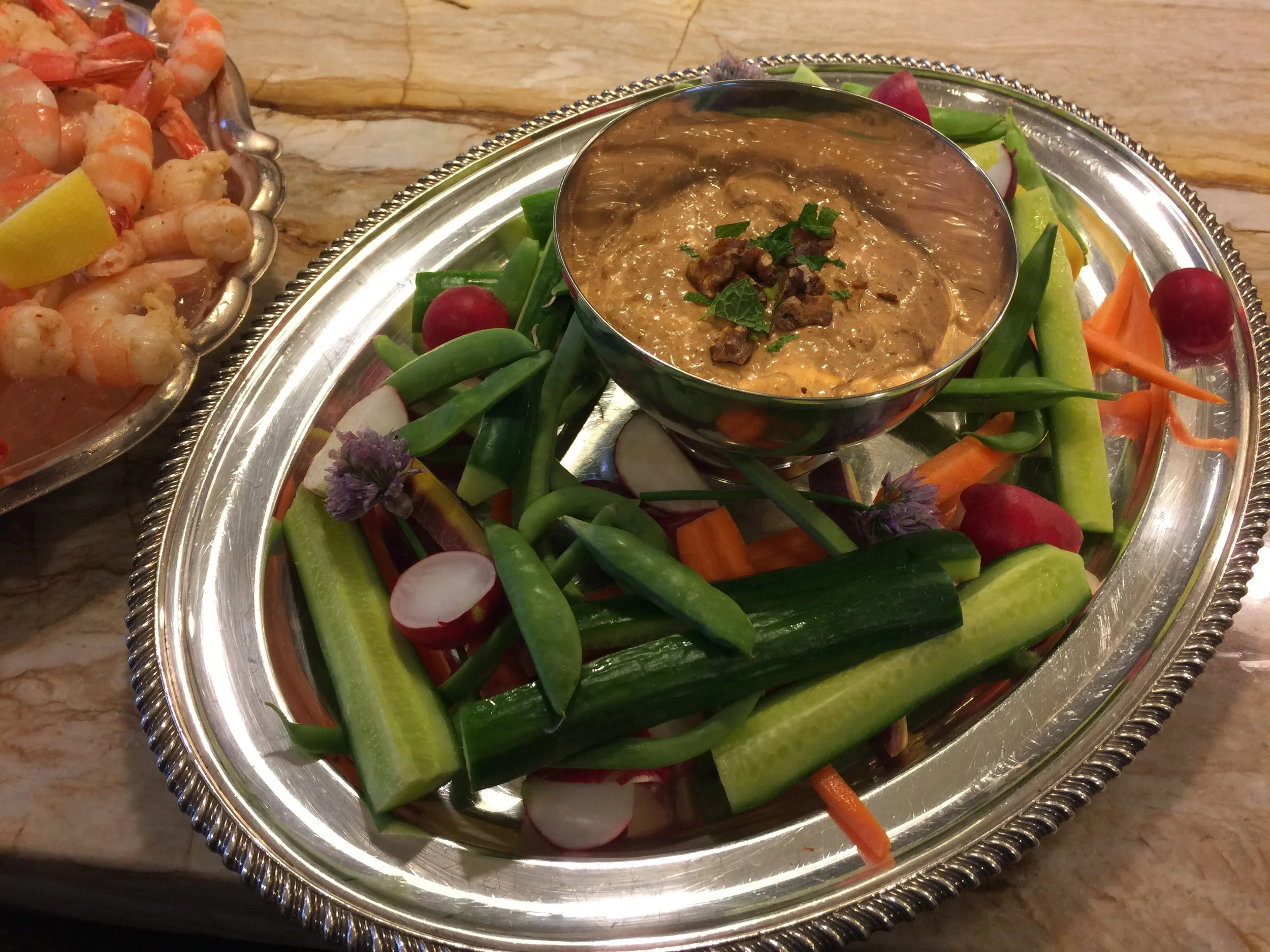 Vegetable platter with cucumber, carrots, radishes, snap peas, and a bowl of creamy dip garnished with herbs, on a silver tray.