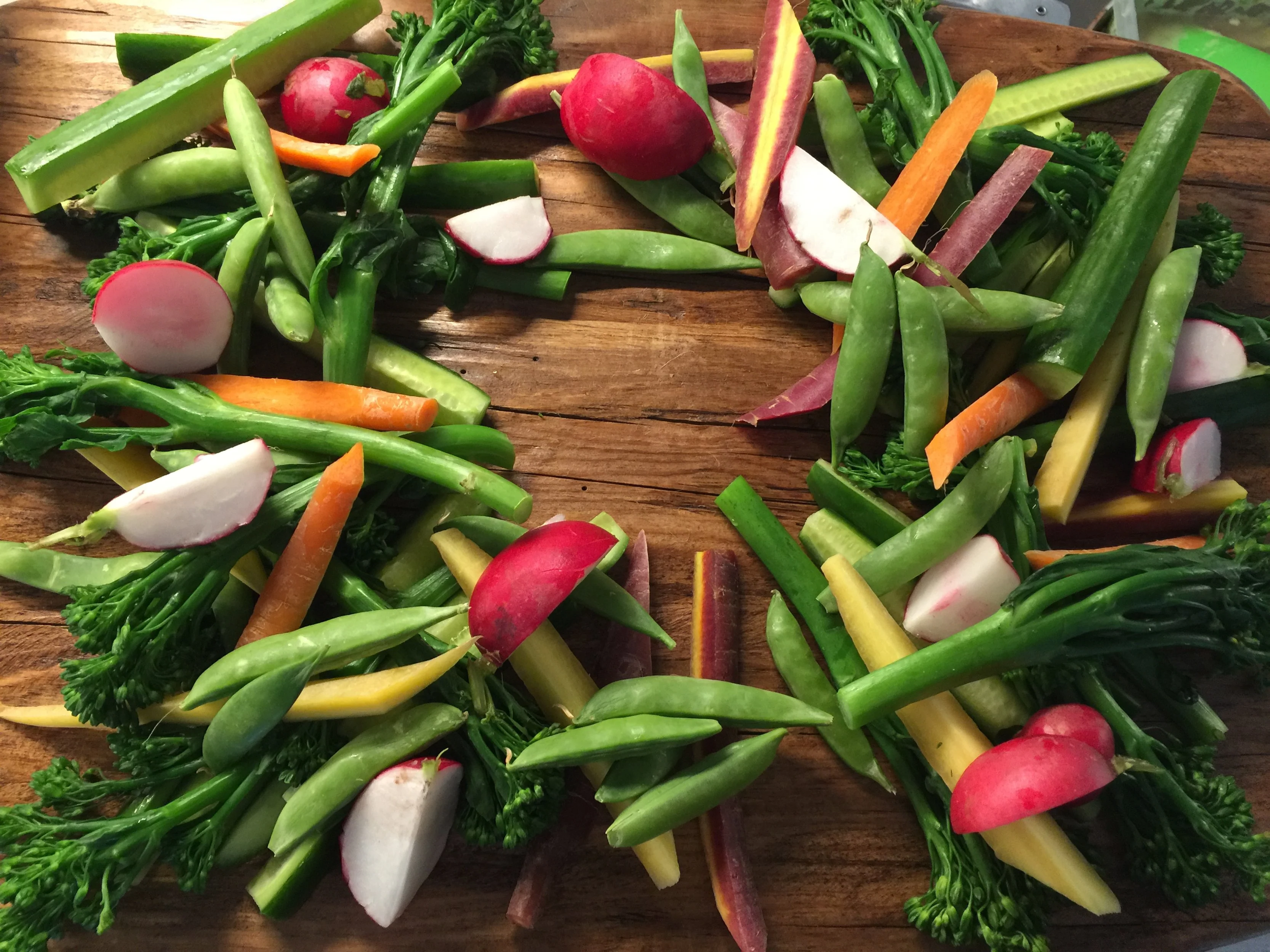 A wooden surface with an arrangement of assorted fresh vegetables, including radish slices, carrot sticks, cucumber pieces, broccoli, snap peas, and leafy greens, arranged in a circular pattern.