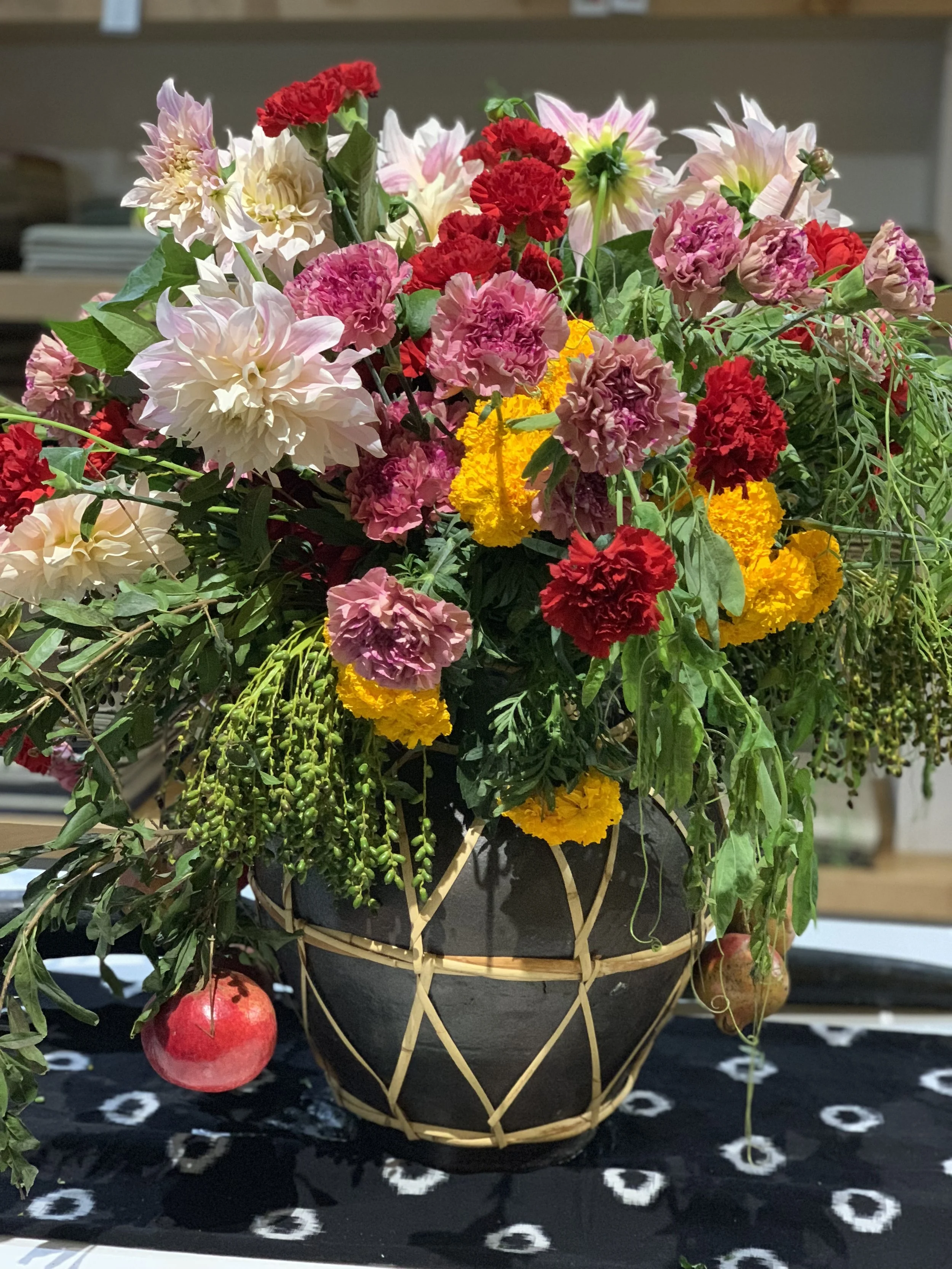 A black and tan decorative basket filled with a variety of colorful flowers, including pink, red, yellow, and white blossoms, sits on a black and white patterned cloth.