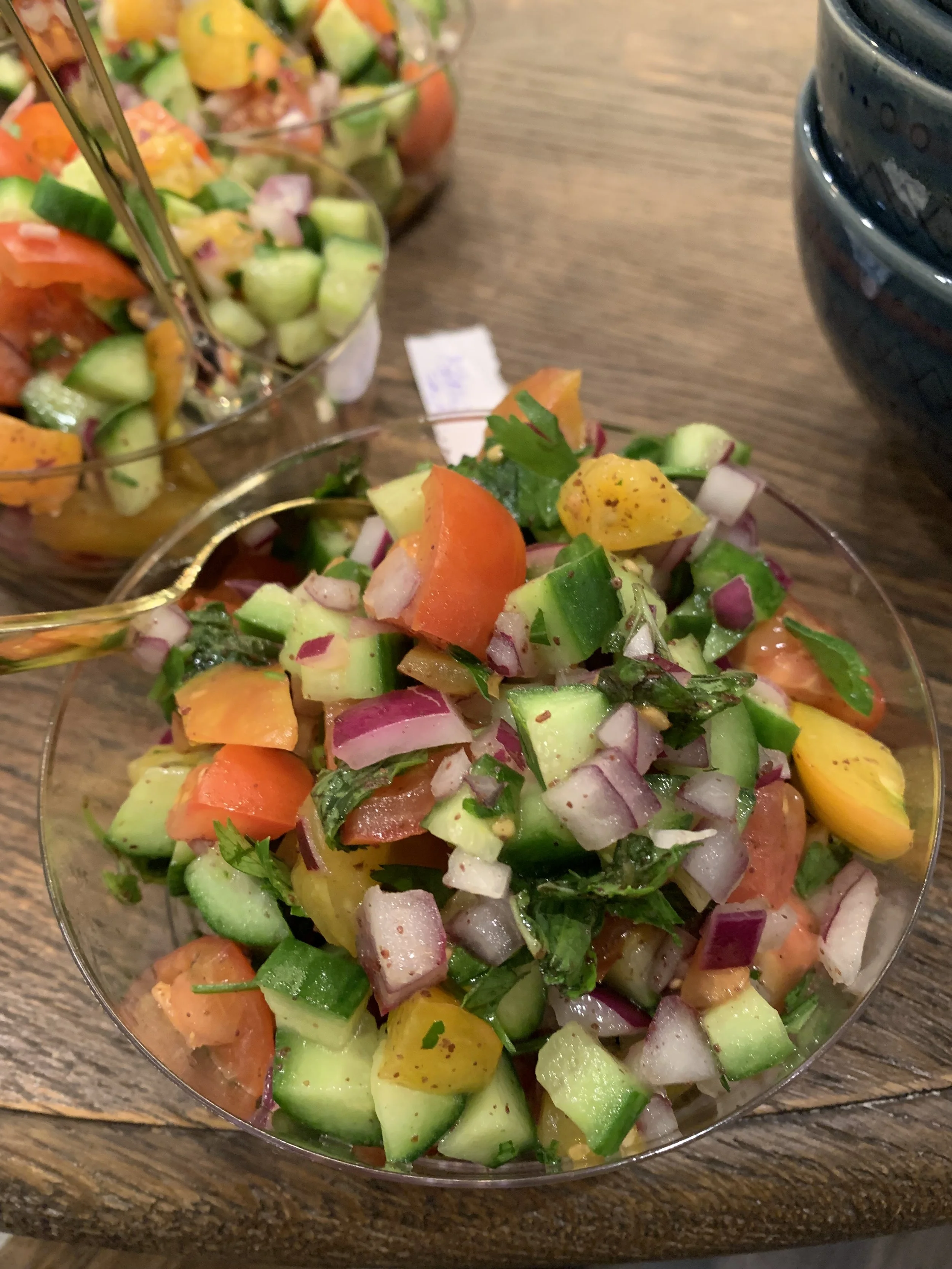 Close-up of a bowl of freshly made cucumber, tomato, onion, and cilantro salad on a wooden table, with additional bowls of salad in the background.