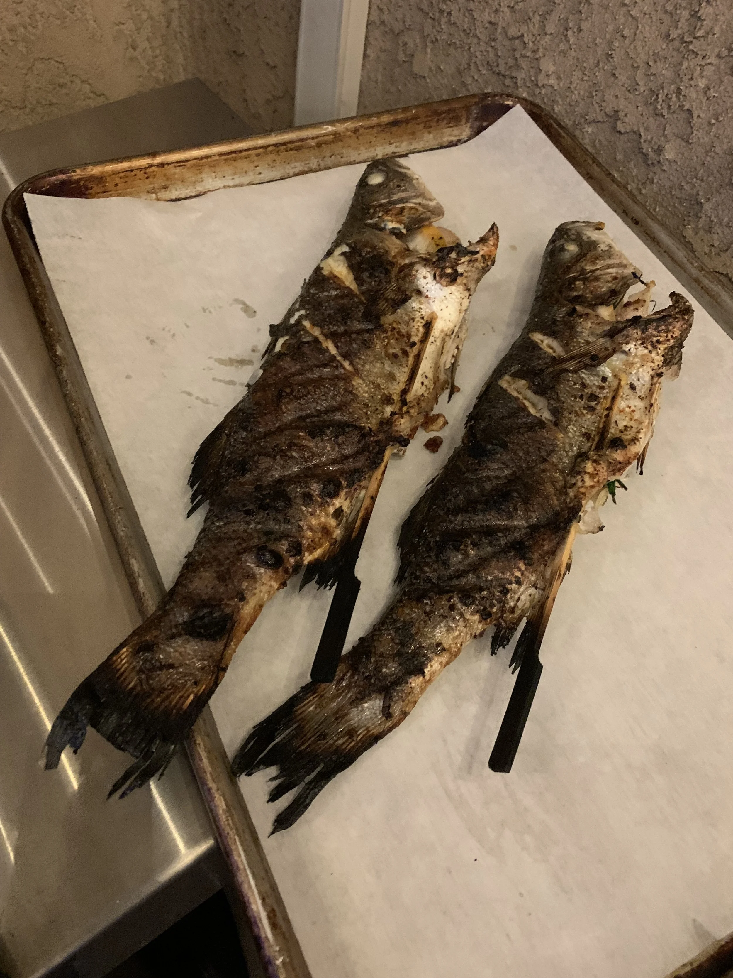 Two grilled whole fish on a baking sheet lined with paper, with a dark crispy skin and visible fins.