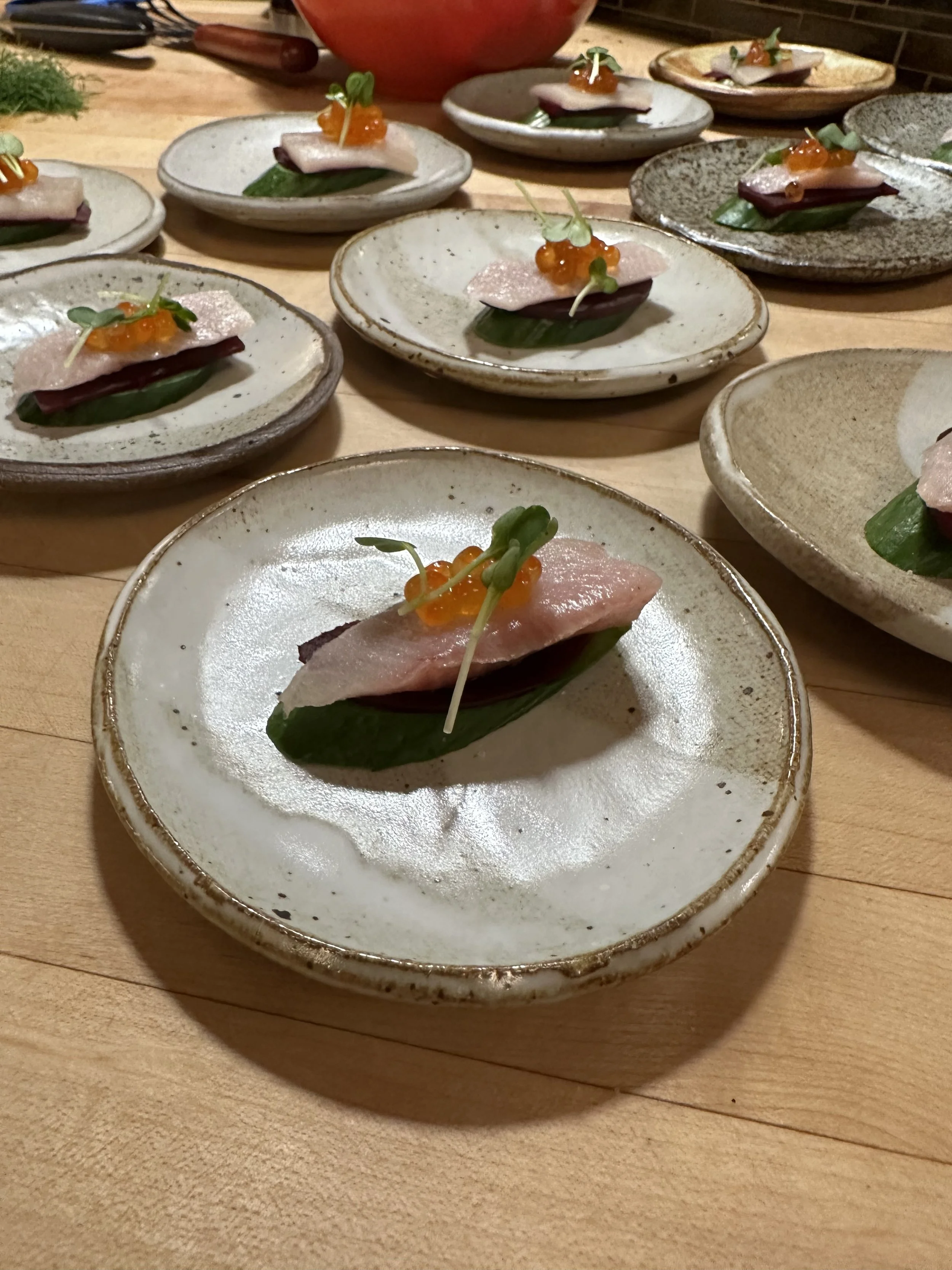 Multiple plates of open-faced sandwiches with a leaf at the base, topped with a slice of meat, orange fish roe, and microgreens, arranged on a wooden table.