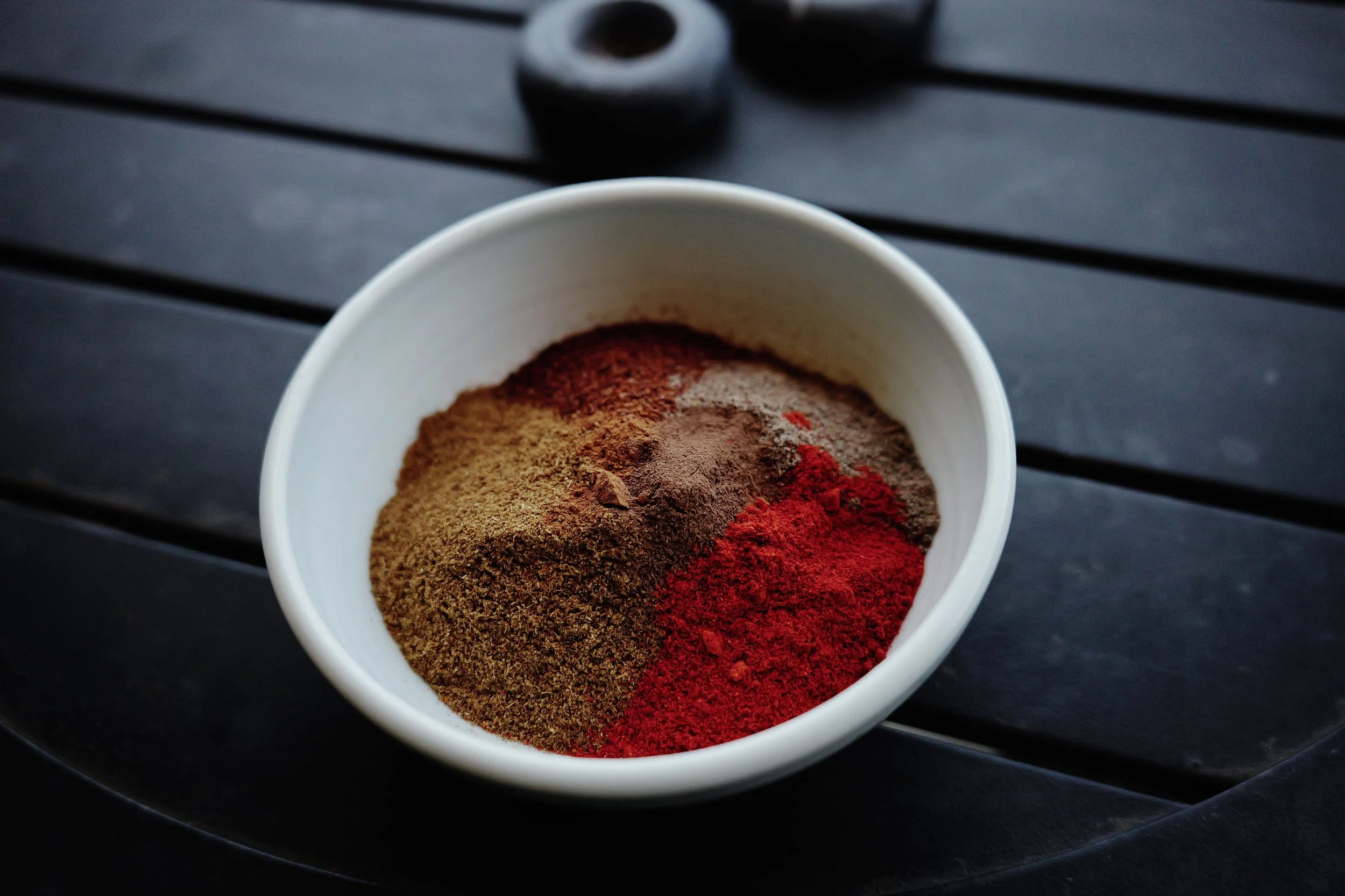 Container of ground spices in various colors including brown, red, and black, placed on a dark wooden surface.