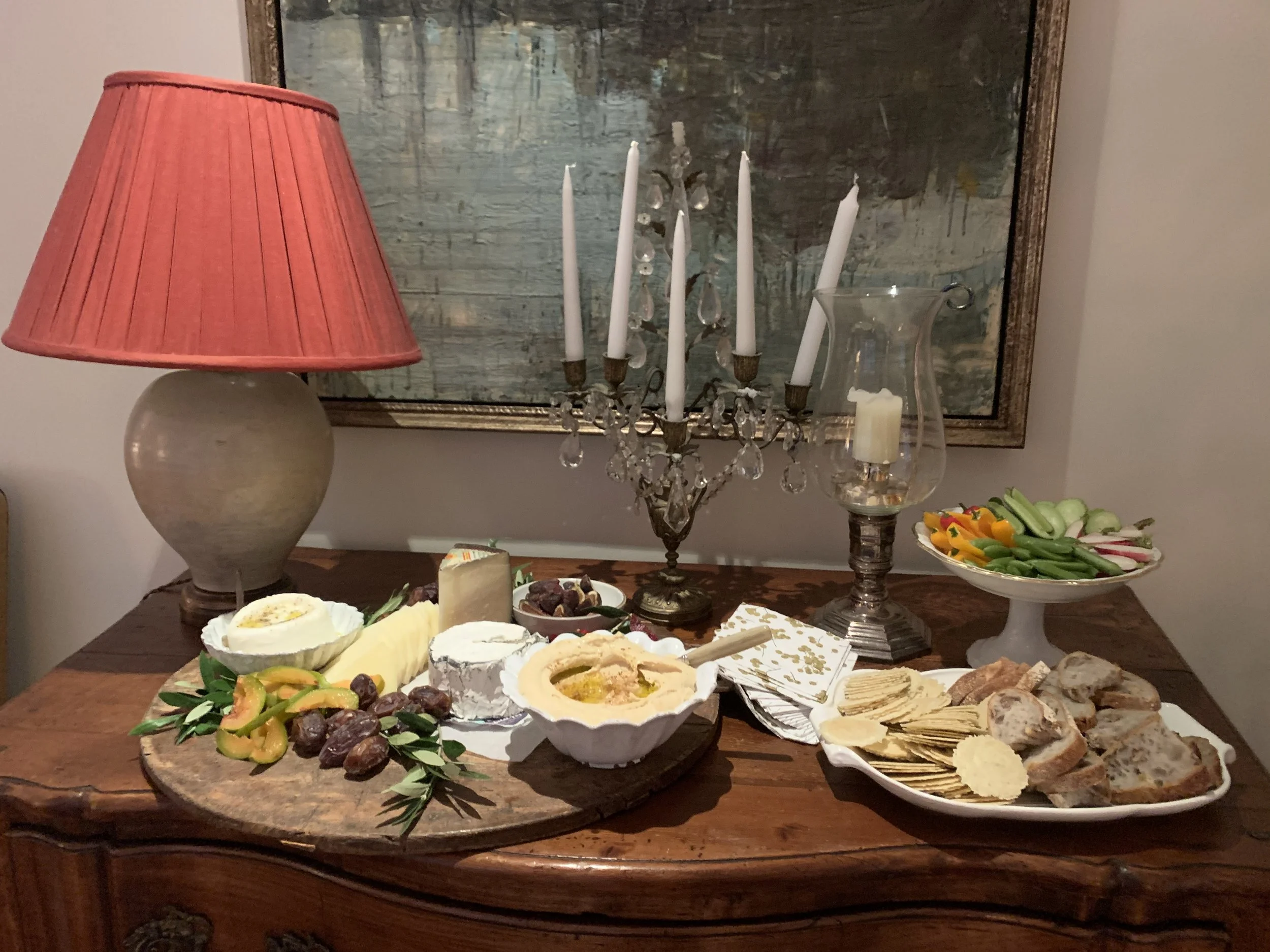 A wooden sideboard with cheese, crackers, vegetables, and a candelabra. A red lampshade on a ceramic lamp and a glass hurricane lamp with a candle. A painting in the background.