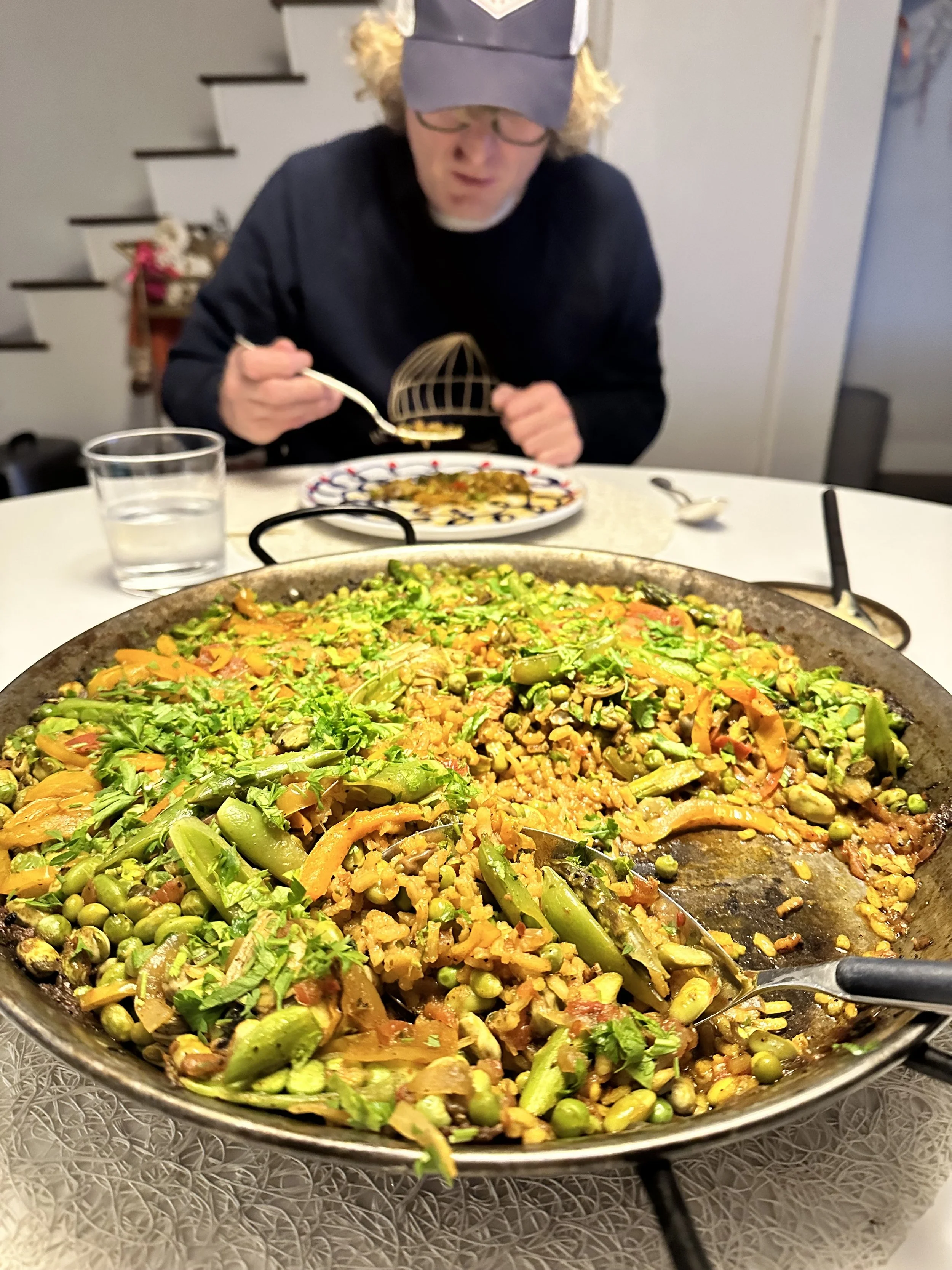 A large skillet filled with a colorful vegetable stir-fry, including peas, carrots, and cabbage, on a table. In the background, a person with glasses, a black sweater, and a gray cap is eating at the table.