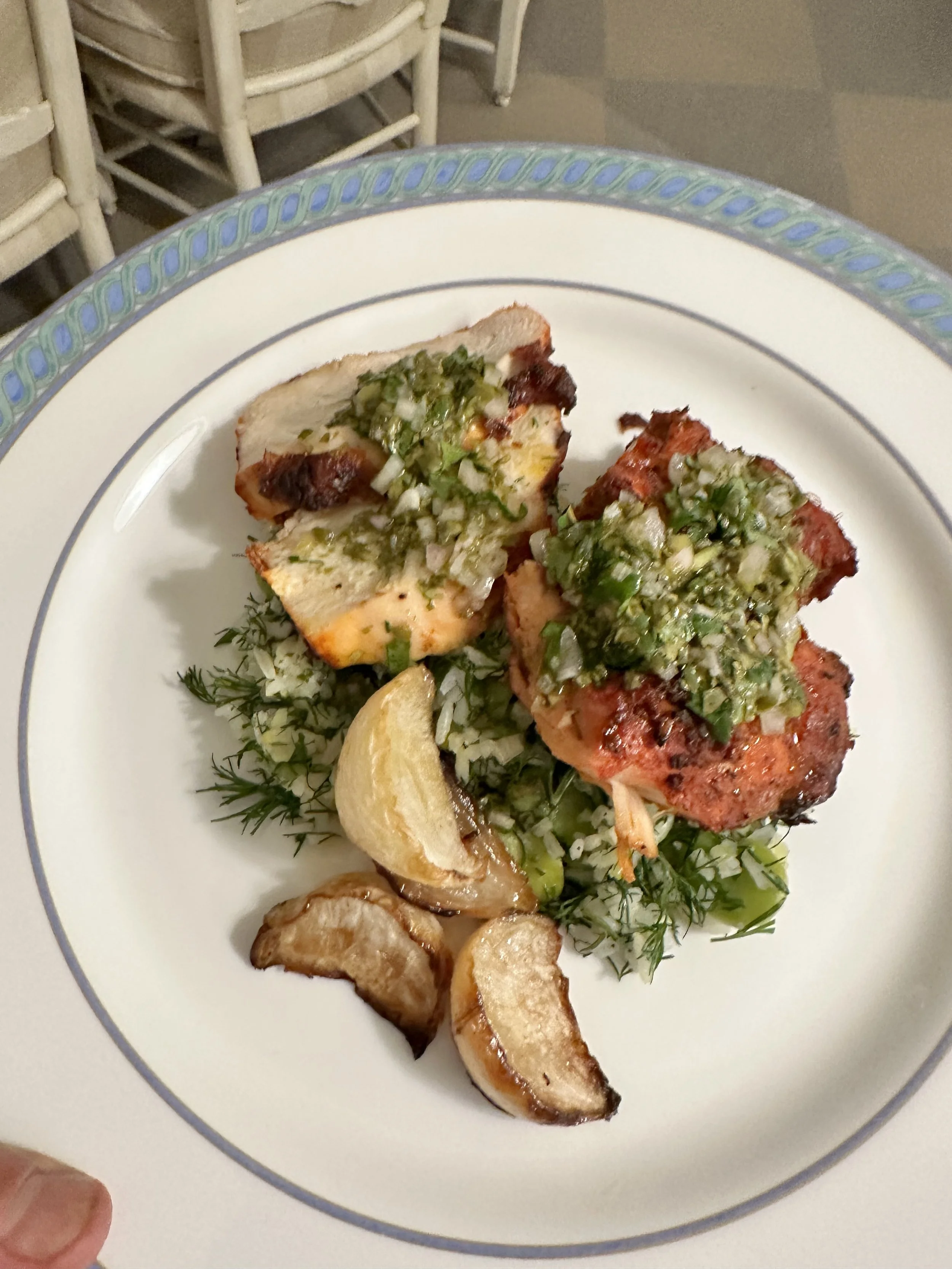 Grilled chicken topped with chopped onions and green herbs, served with roasted potatoes and a lemon wedge on a decorative white plate.