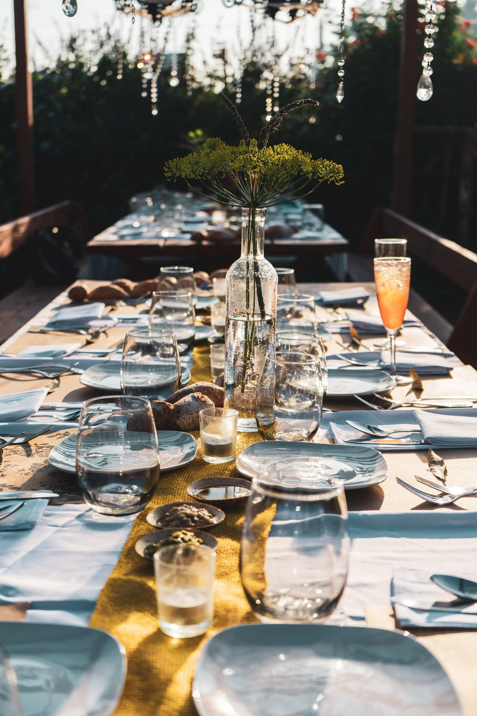 A table set for outdoor dining with plates, glasses, silverware, and a centerpiece of flowers in a glass bottle, illuminated by natural sunlight.