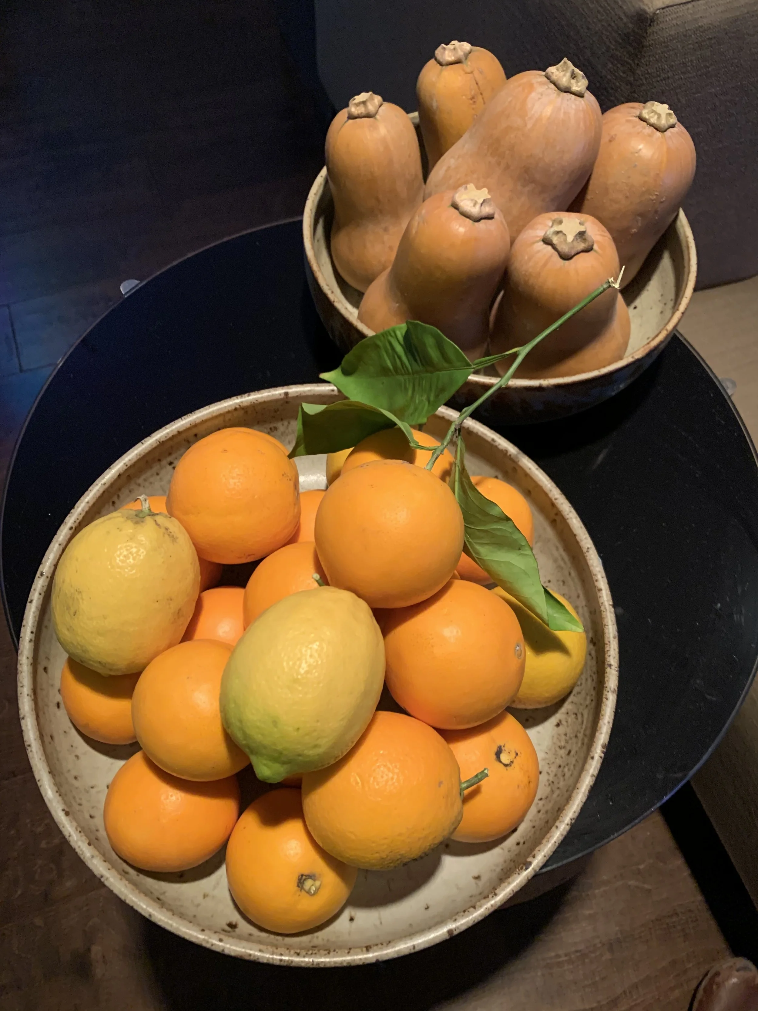 Two ceramic bowls filled with fresh fruits, one with oranges and lemons in the foreground, and the other with butternut squash in the background on a black table.