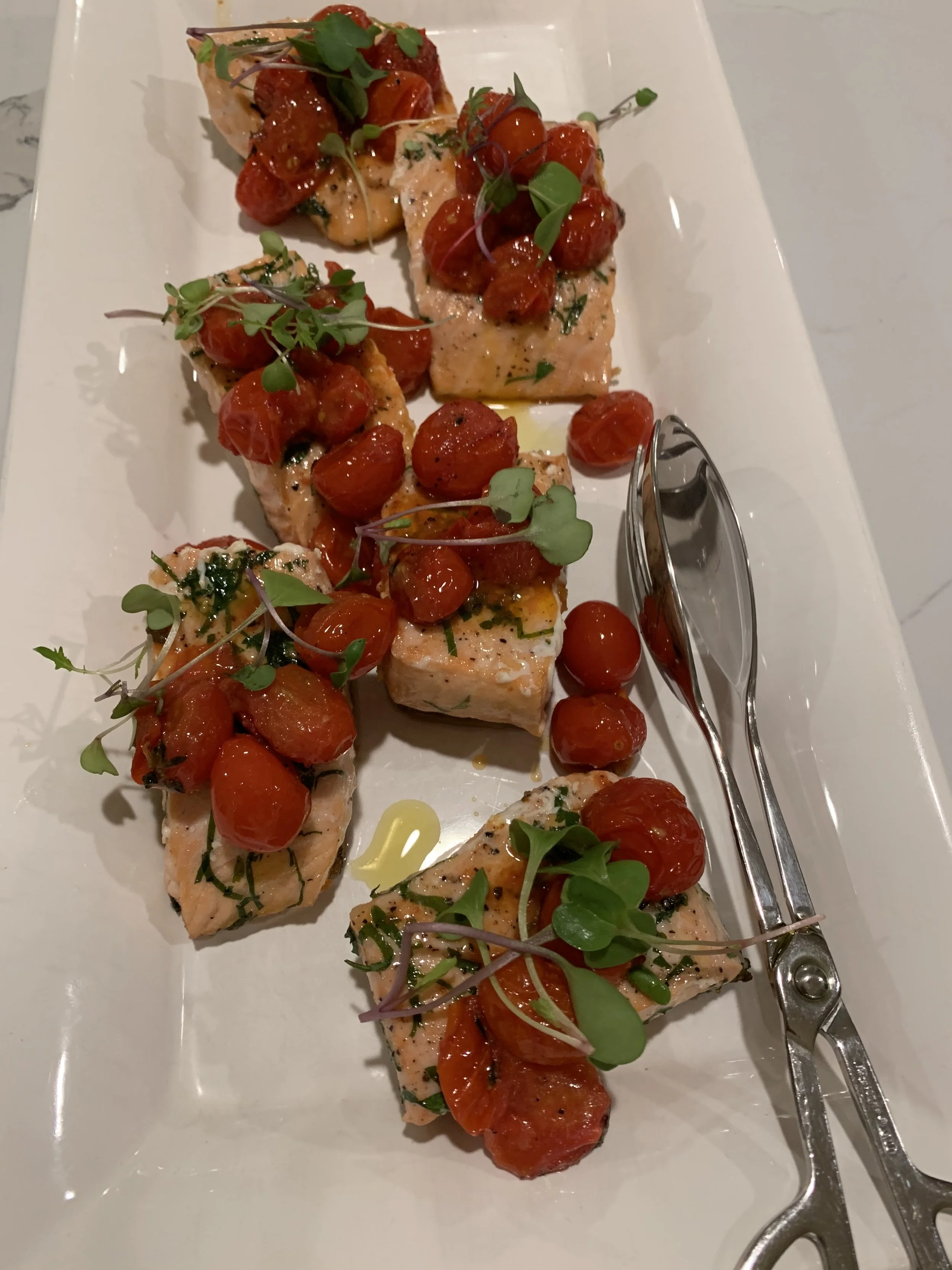Salmon bites topped with cherry tomatoes and microgreens on a white rectangular plate, with a pair of scissors placed on the side.