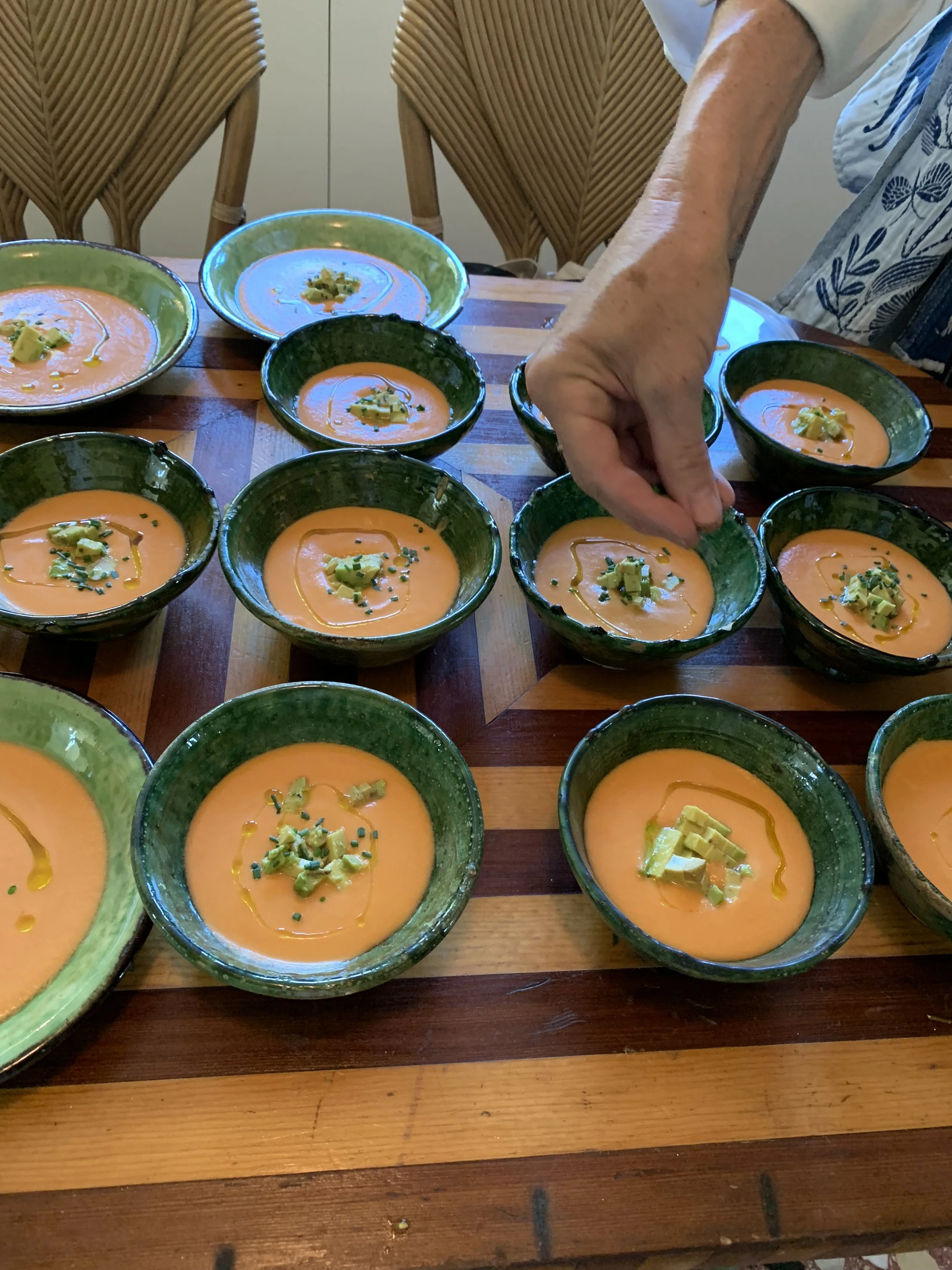 Multiple bowls of orange soup garnished with chopped green herbs and a drizzle of oil on a wooden table, with a person's hand reaching to touch one bowl.