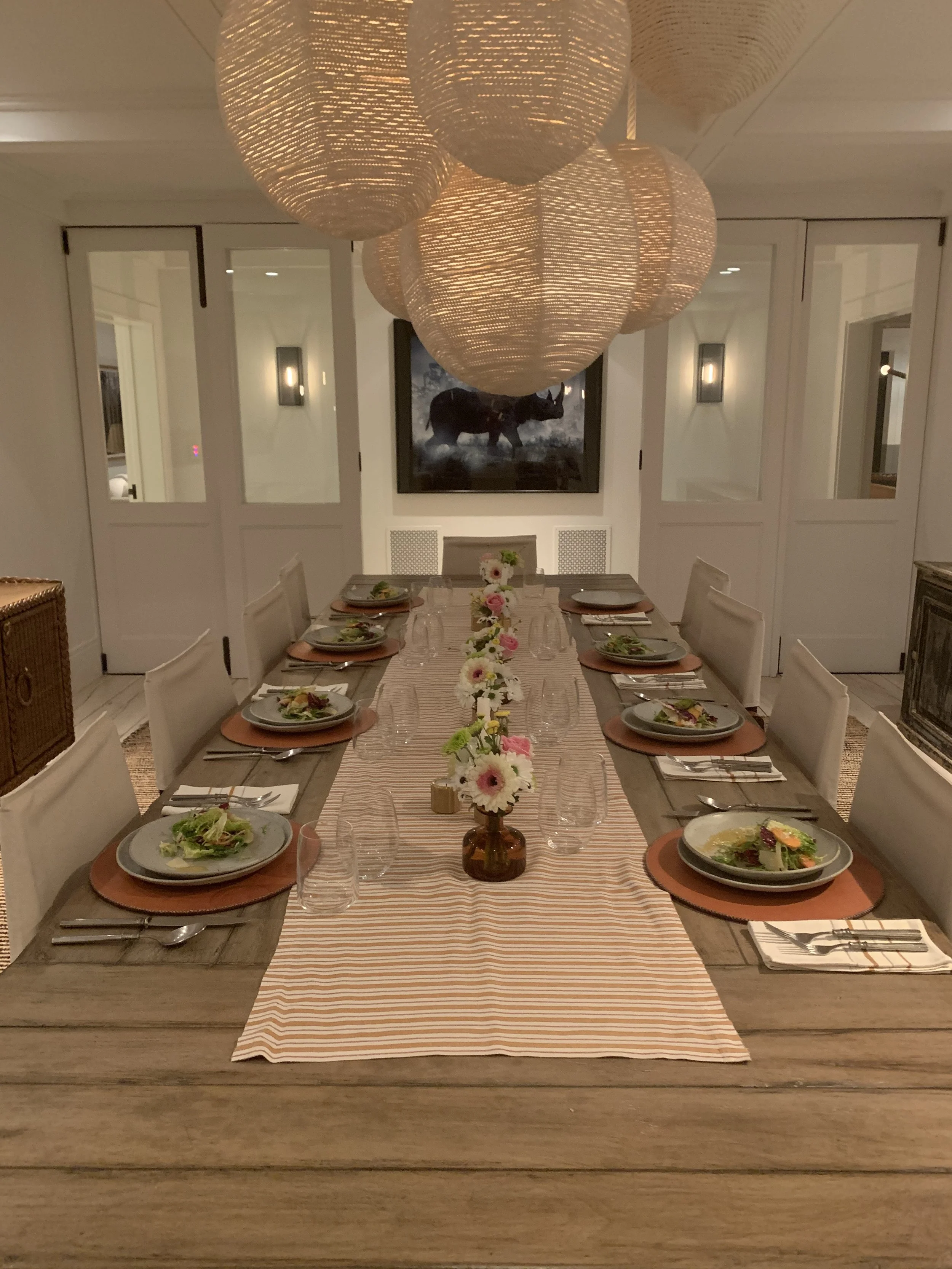 A dinner table set for eight, with plates of salad, wine glasses, and floral centerpieces, under large woven pendant lights in a dining room with white walls and artwork.