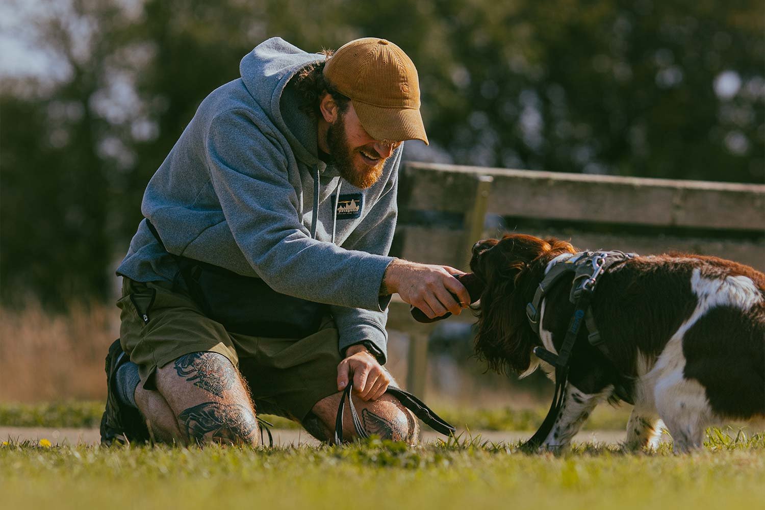 A man with a beard, wearing a beige cap, grey hoodie, and camo shorts with tattoos, is kneeling on the grass and playing with a black and white dog, both smiling and engaging in a friendly interaction outdoors.