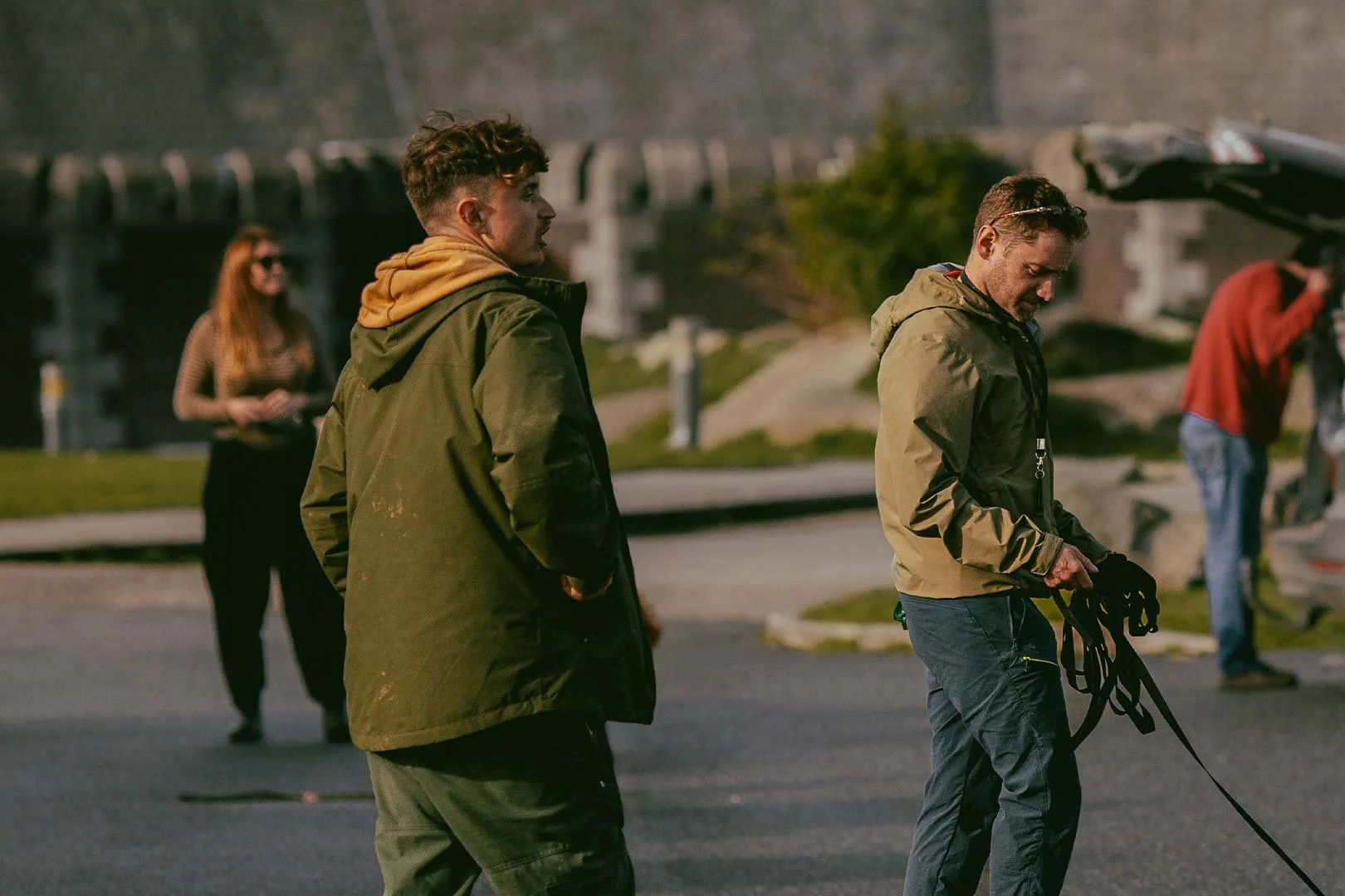 Two young men in green jackets standing on a street, one looking down and holding a leash, while the other looks ahead. In the background, a woman in sunglasses and a man in a red hoodie are also present.