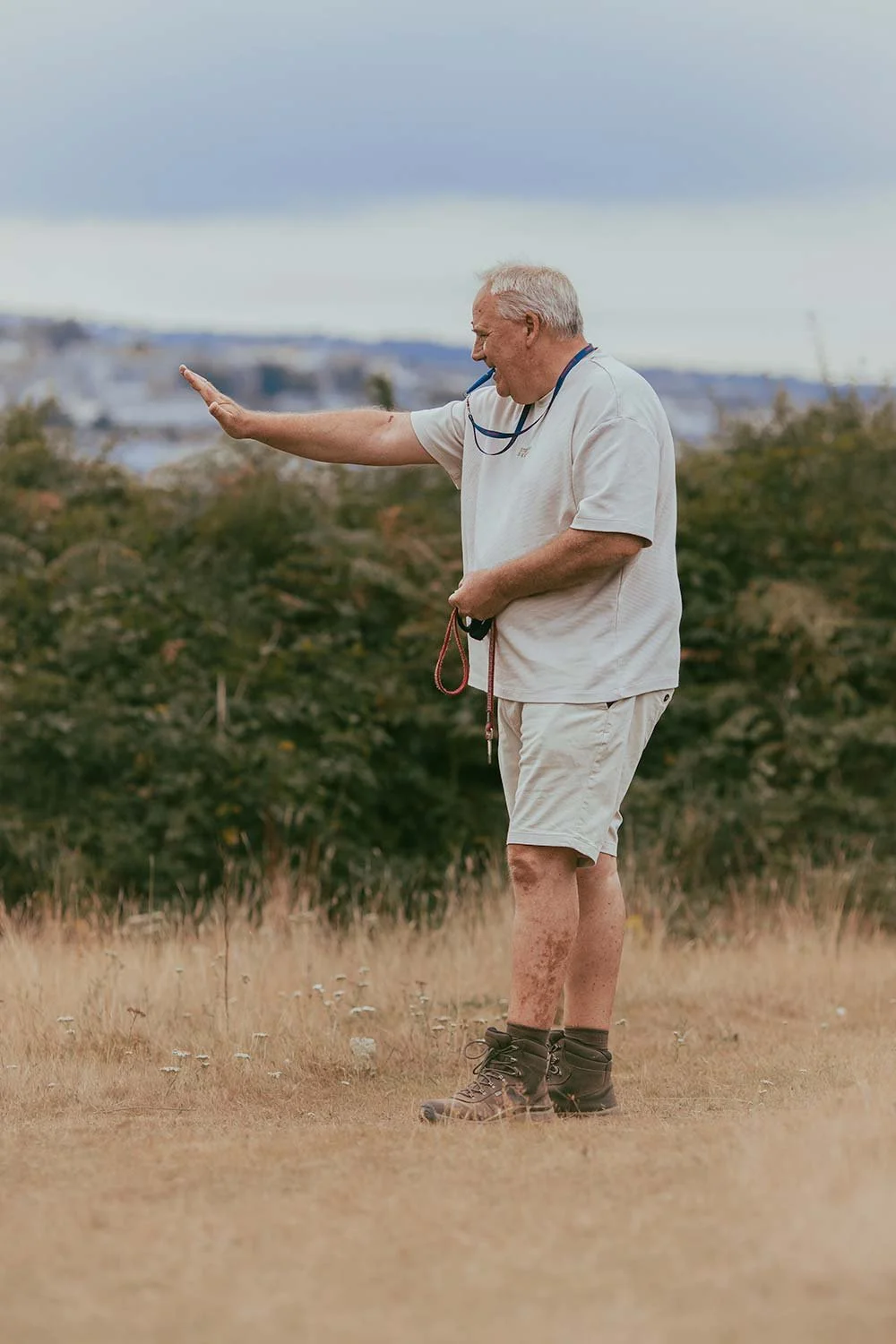 An elderly man with white hair and a white T-shirt stands outdoors on a grassy field, smiling and waving with his right hand while holding sunglasses in his left hand, with mountains or a city skyline in the distance under a cloudy sky.