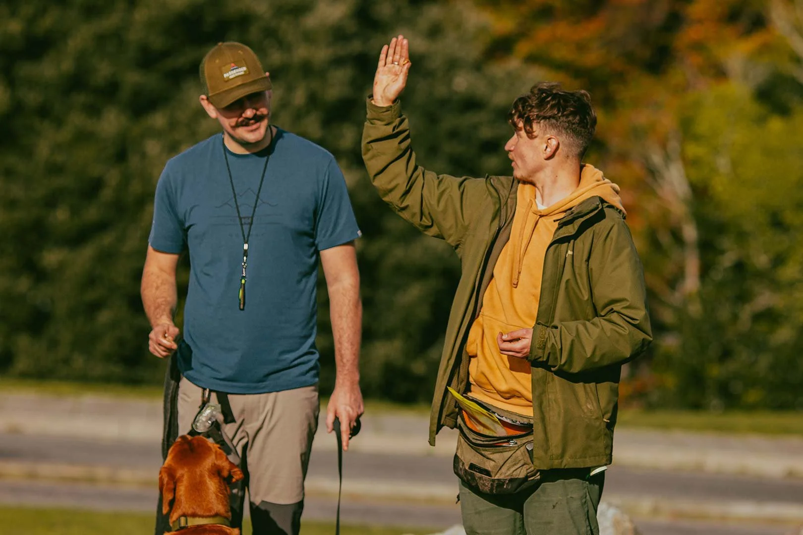 Two men walking outdoors, one raising his hand for a high-five, with a dog on a leash, trees in the background.