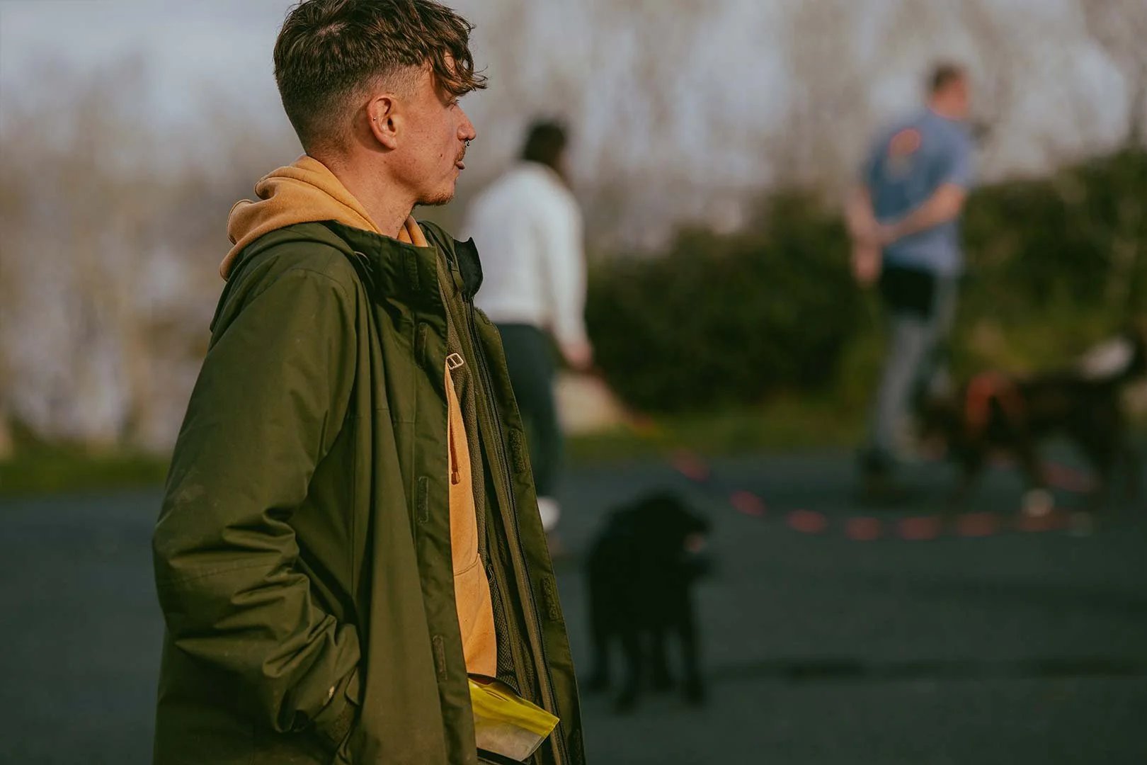 A young man with brown hair wearing a green jacket and yellow hoodie standing in profile outdoors, with a blurred background of people and trees.