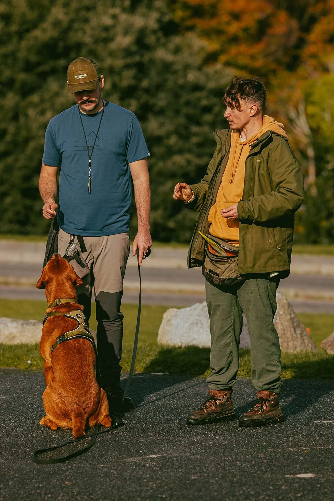 Two men and a brown dog outdoors with trees in the background.