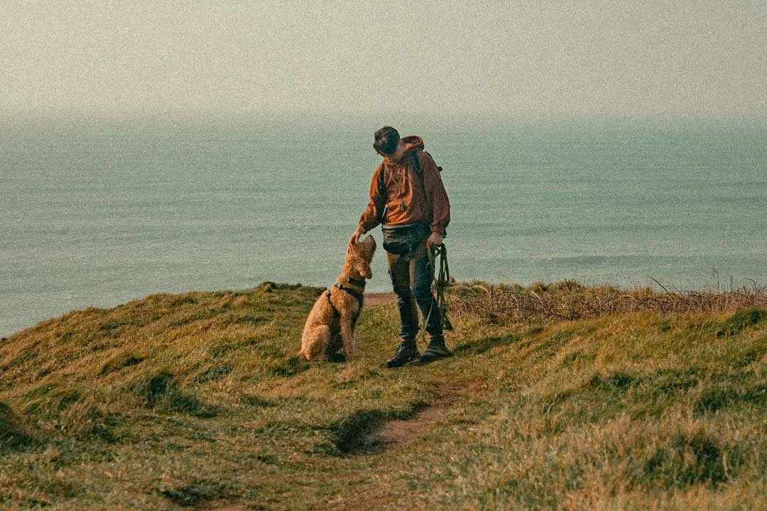 Elliot and Merryn stood on a clifftop at Poldhu Cove, he's stroking Merryn on her head