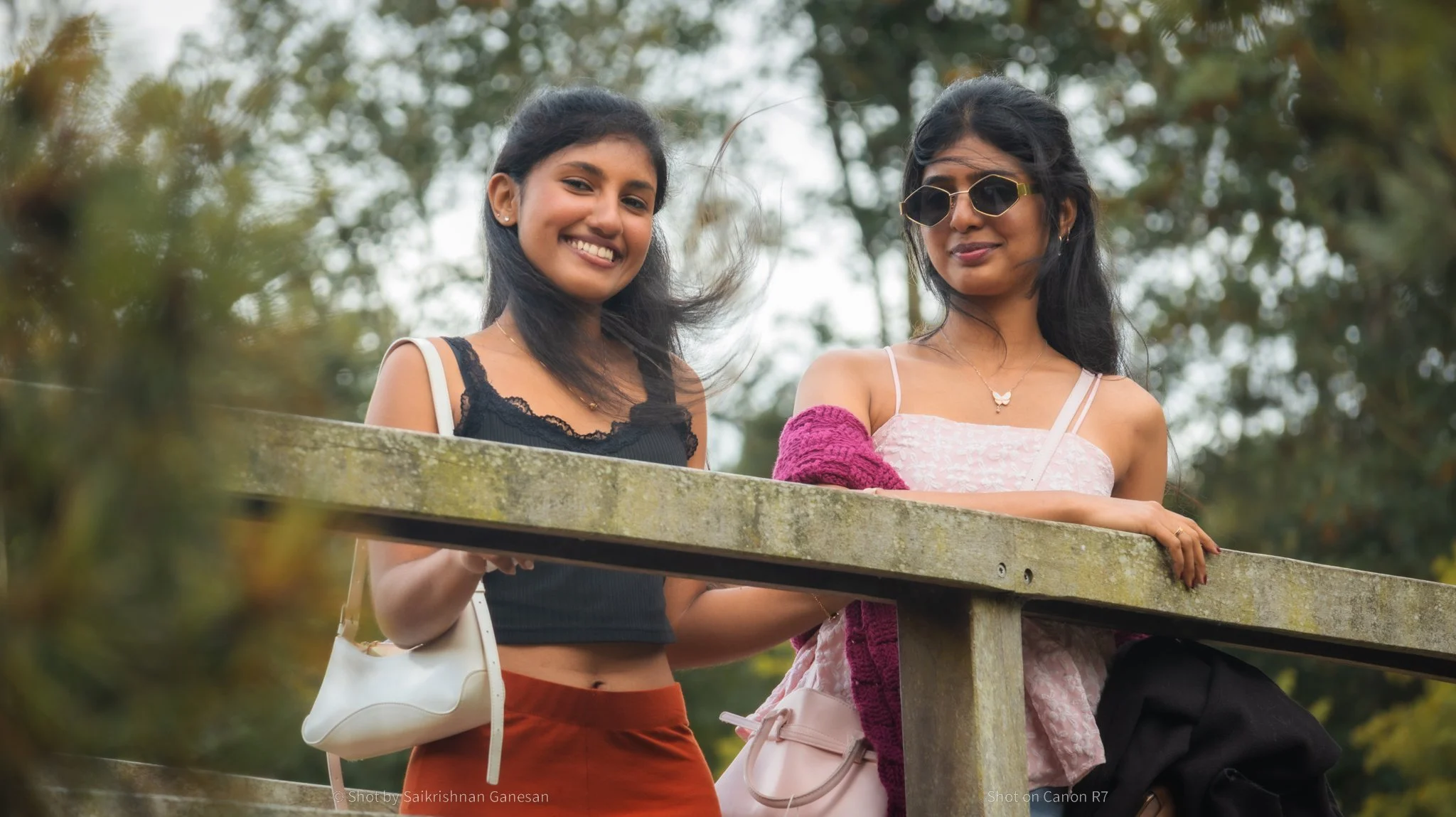Two young women standing outdoors near a concrete railing with trees and cloudy sky in the background. One woman is smiling, wearing a black top with lace details, a small white handbag, and red pants. The other woman is wearing sunglasses, a pink dress with thin straps, and has a pink sweater draped over her arm.
