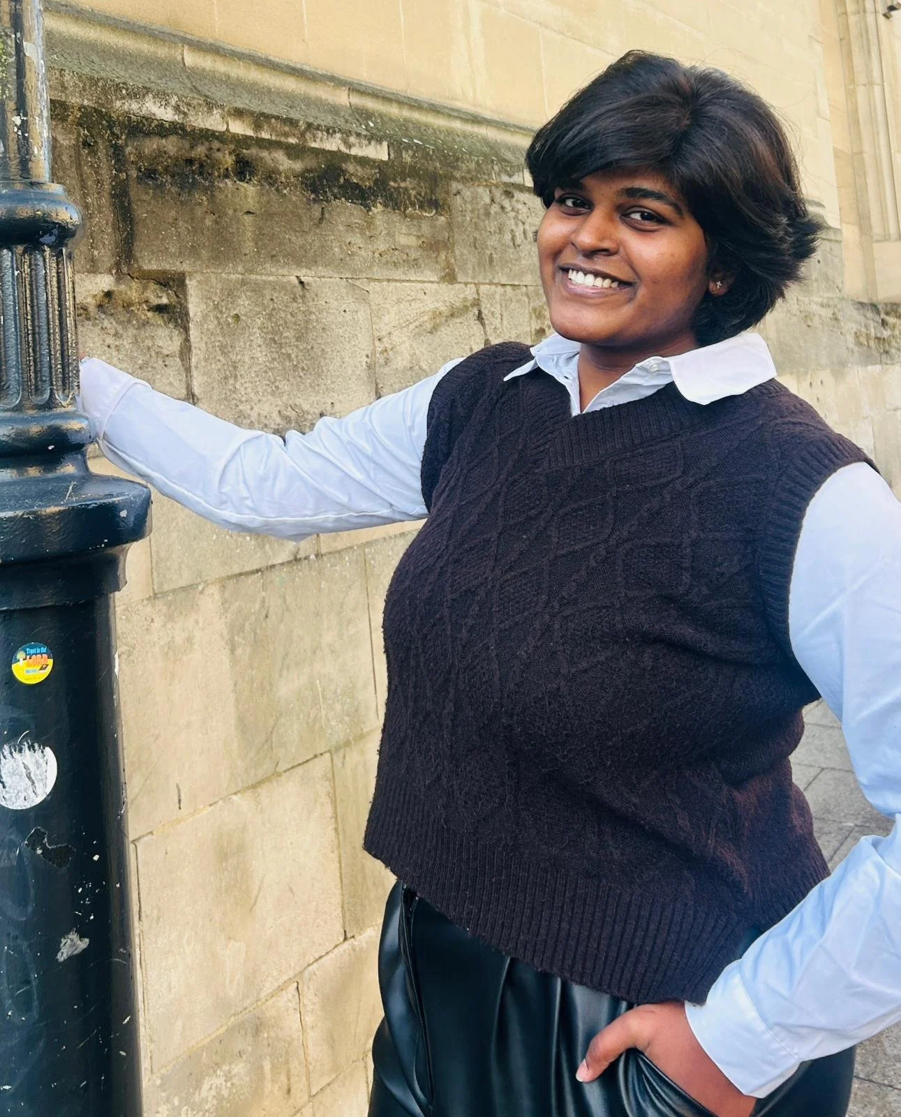 A woman smiling, wearing a white shirt with a dark sweater vest and leather pants, standing outdoors beside a stone wall and a black utility pole.