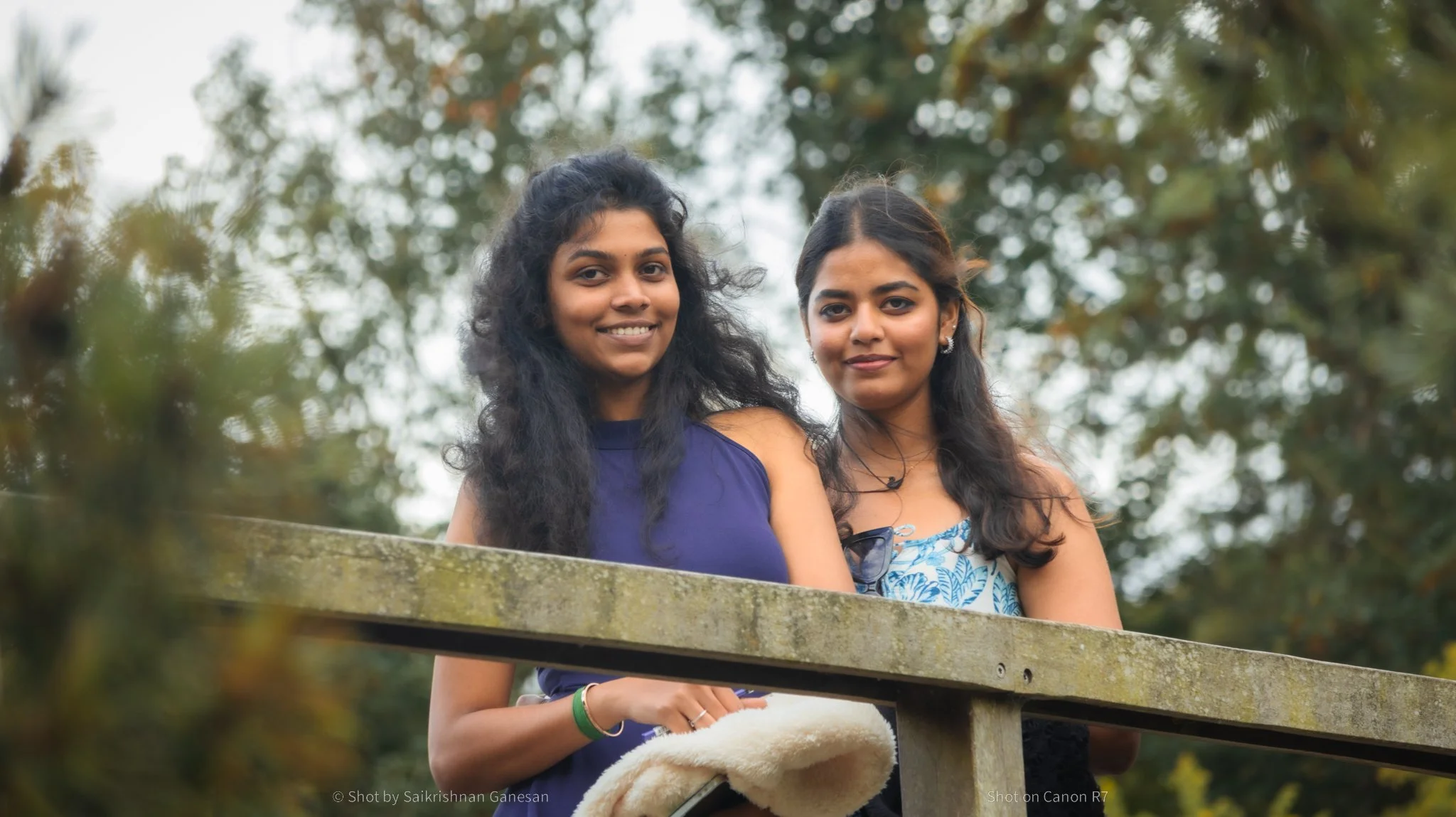 Two young women standing outdoors behind a wooden railing, smiling at the camera with a background of trees and cloudy sky.