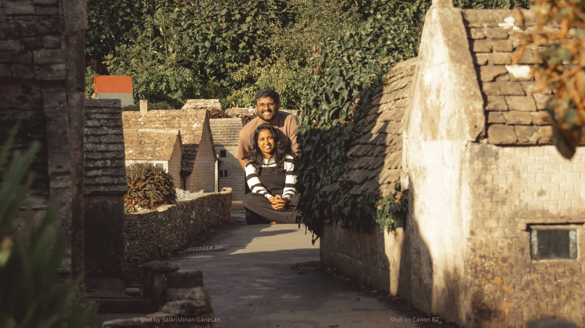 A woman sitting cross-legged on the ground, smiling, with a man standing behind her in a small village with old stone houses and greenery.