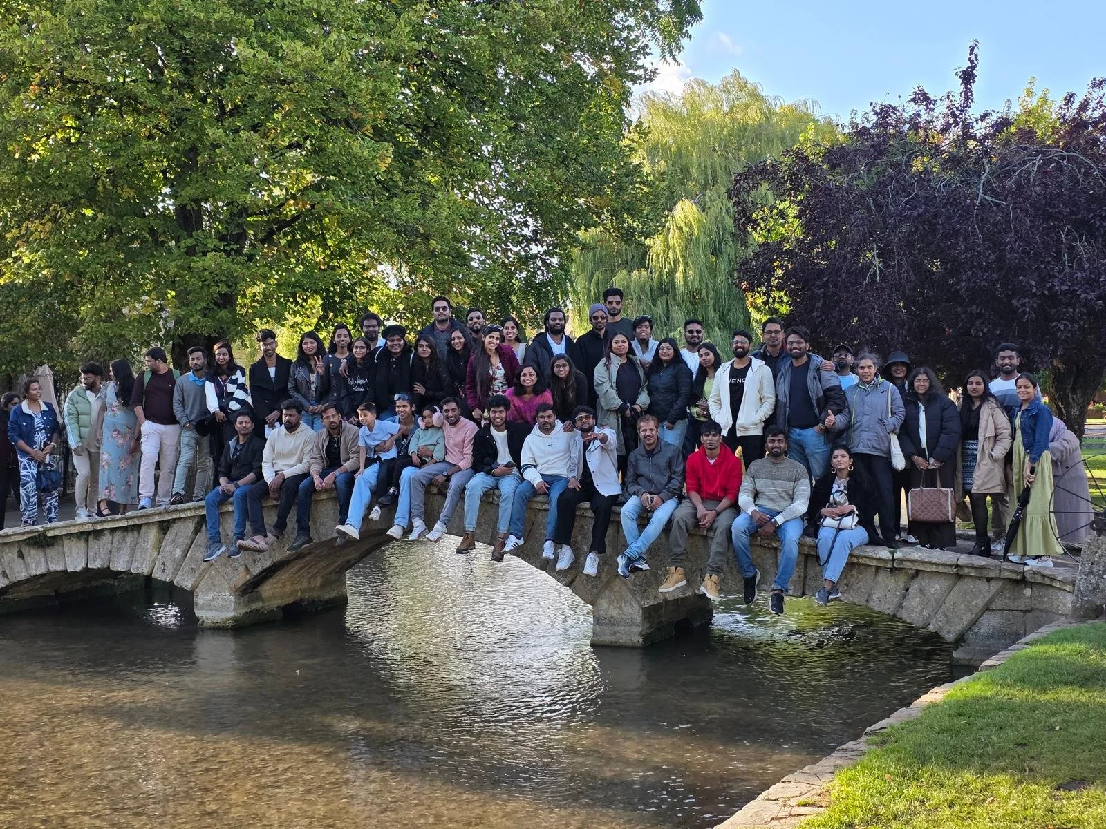 A large group of diverse people pose on a stone bridge over a small river in a park with lush green trees in the background.