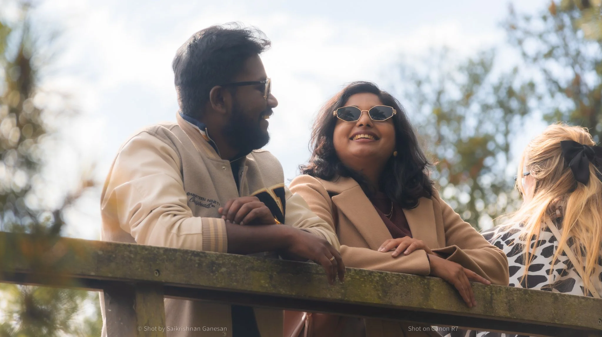 Three people, two women and one man, standing outdoors on a balcony or railing during daytime. They are smiling and wearing sunglasses, with trees and a cloudy sky in the background.