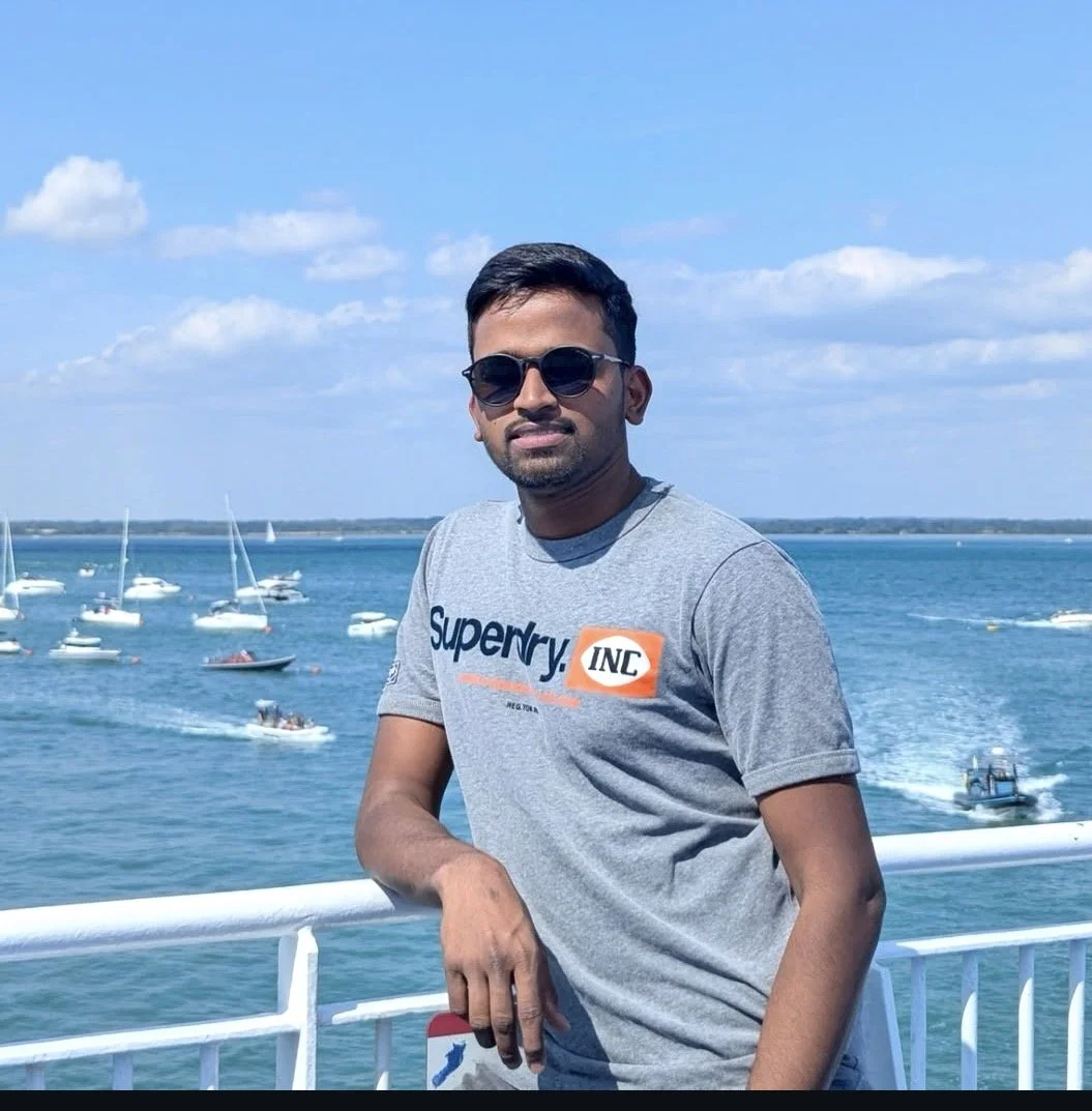 A man wearing sunglasses and a gray Superdry t-shirt stands by a white railing near a body of water with sailboats and small boats, under a blue sky with some clouds.