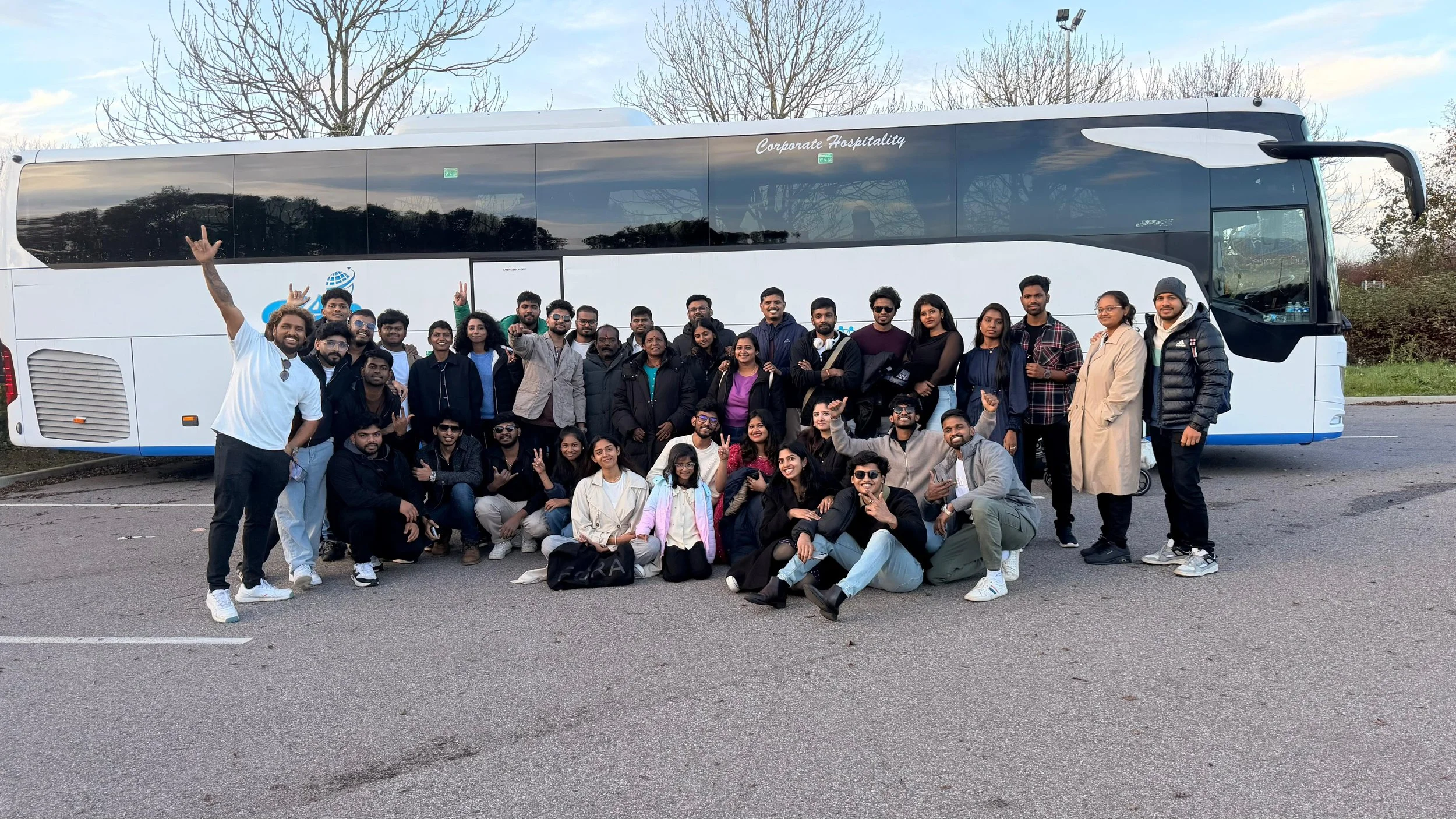 Group of people posing in front of a large white tour bus parked on an open road with trees in the background.