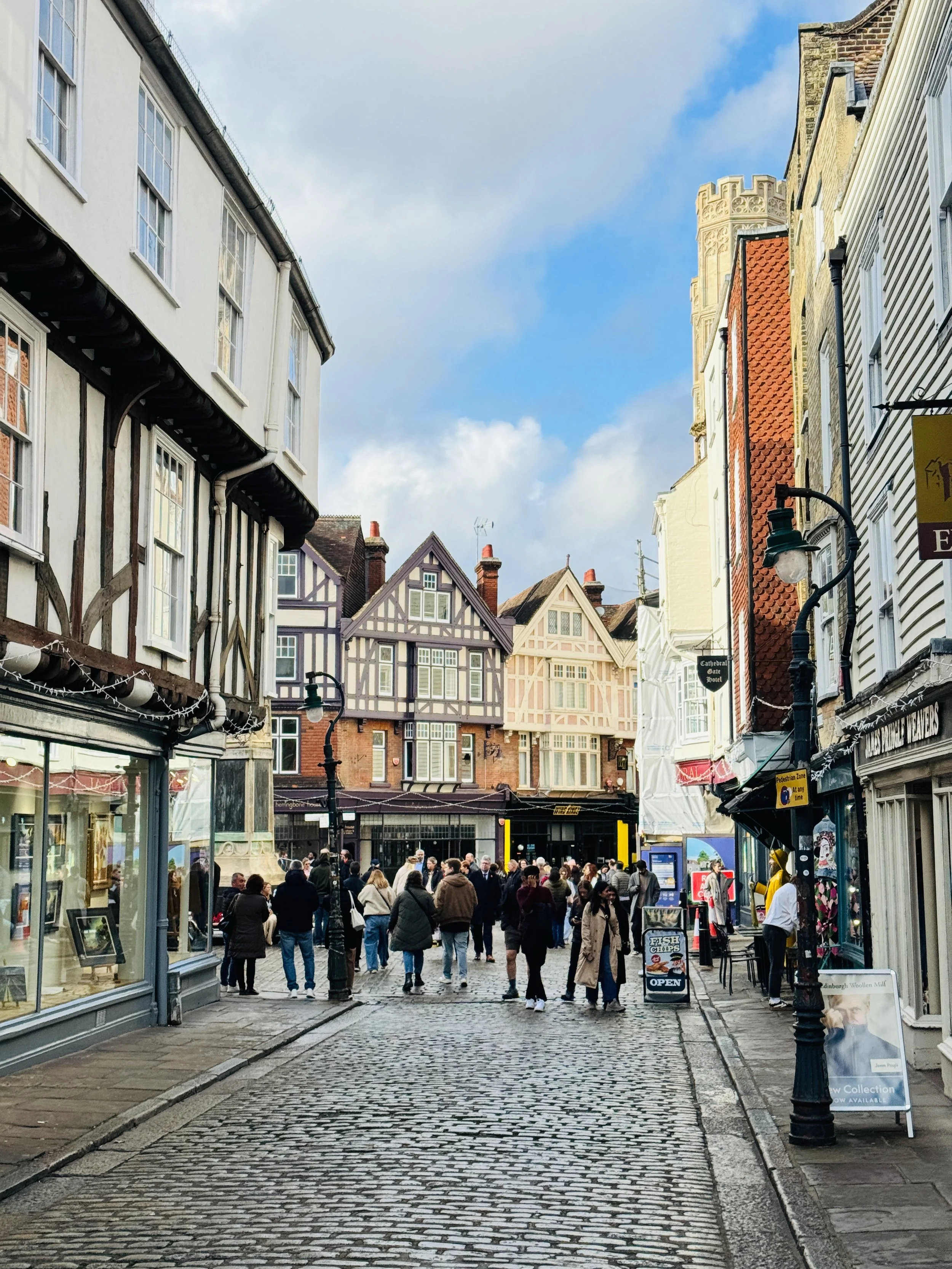People walking on a cobblestone street lined with historic buildings with colorful timber frames and shops on a sunny day.