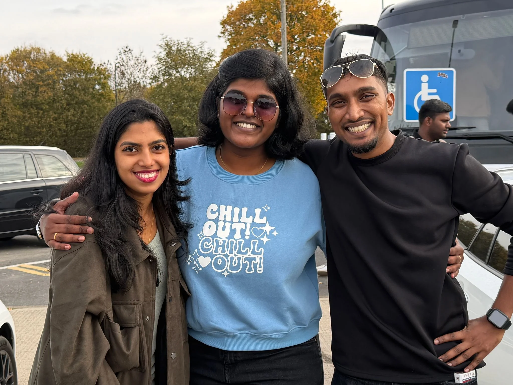 Three friends smiling and posing together outdoors in a parking lot with trees in the background, one wearing a blue "Chill Out!" sweatshirt, another in a black top, and the third in a brown jacket.