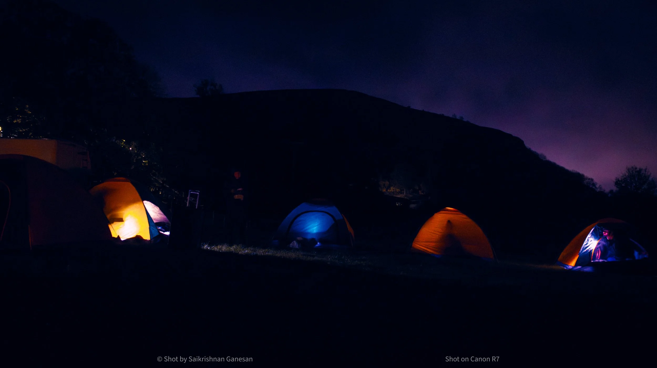 Nighttime scene of four illuminated camping tents set up on grassy terrain, with a dark hillside or mountain in the background and a purple sky overhead.