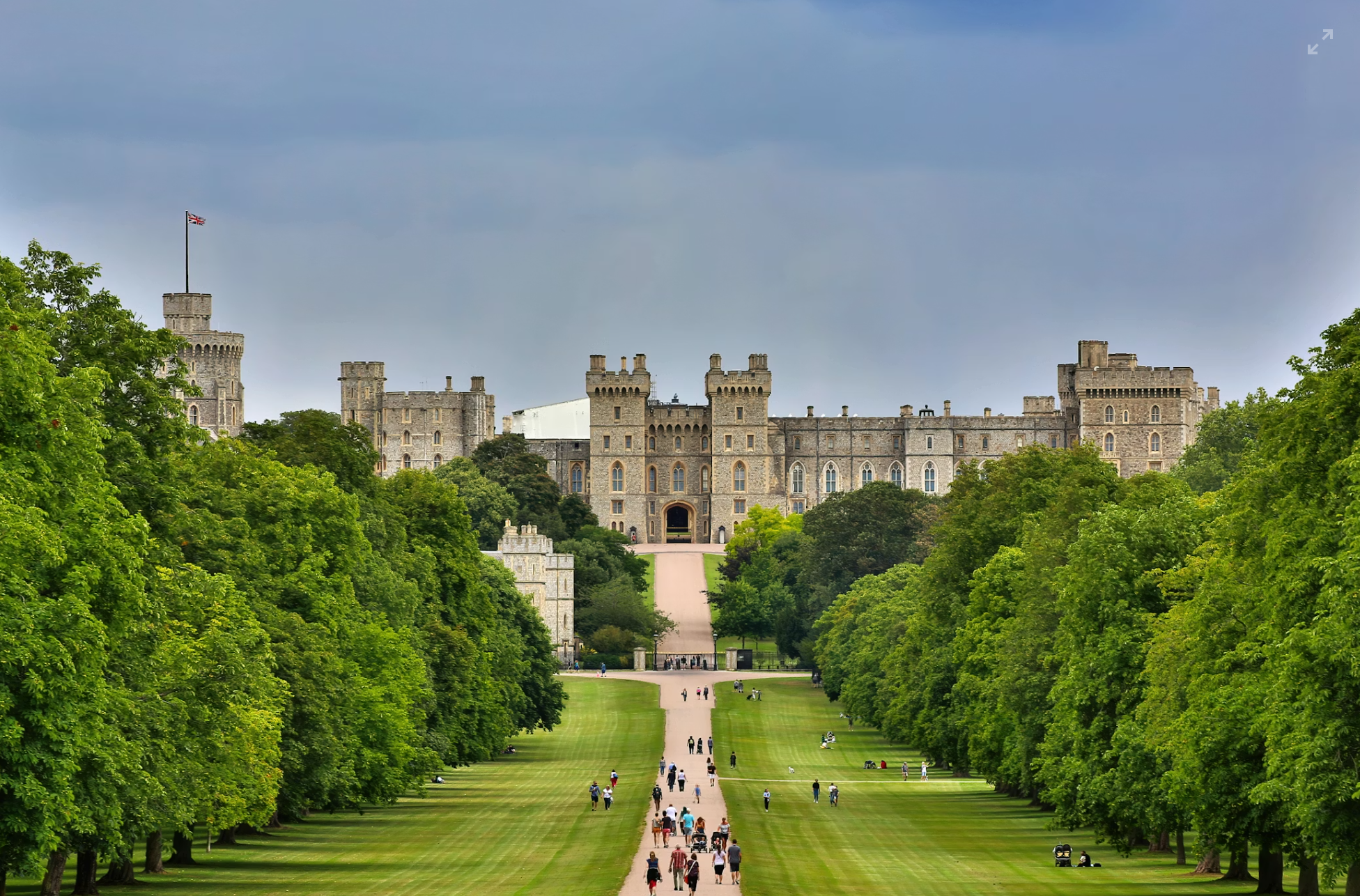 Stone castle with multiple towers and a flag on top, surrounded by lush green trees, with a wide pathway leading up to it and people walking and relaxing on the lawn.