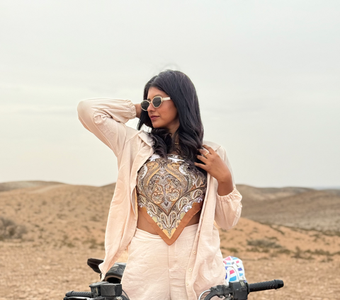 Woman with long dark hair wearing sunglasses, a beige jacket, and a patterned top, standing in a desert landscape with mountains in the background.