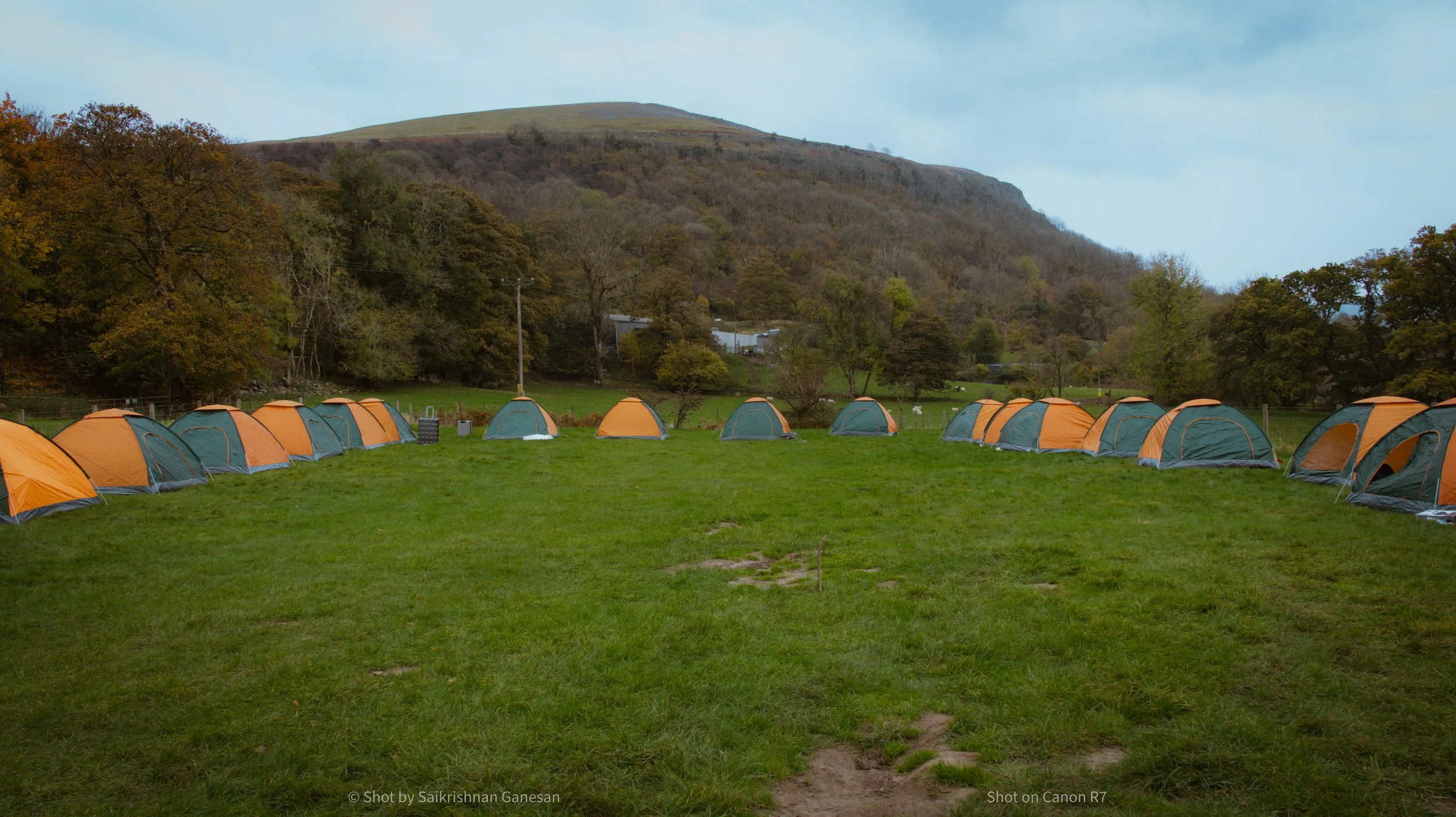 A campsite with multiple orange and green tents pitched on a grassy field surrounded by trees and hills.