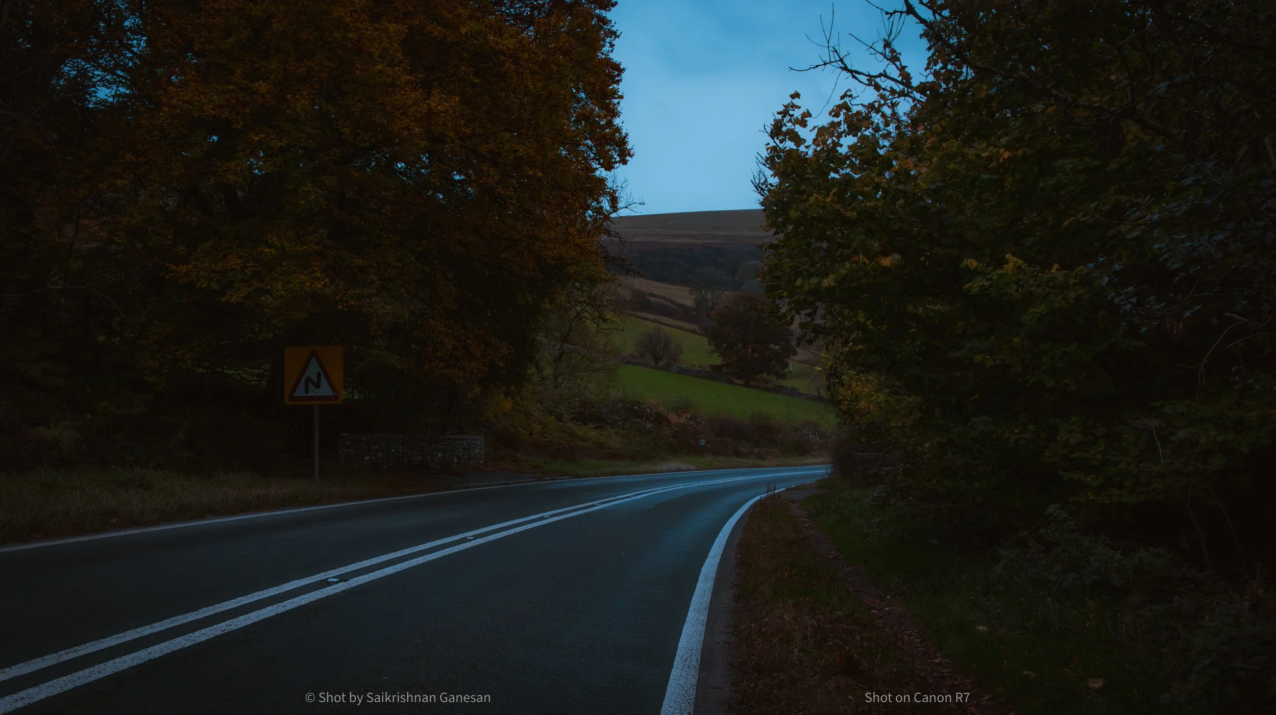A scenic rural road winding through hilly landscape with trees on both sides during dusk or dawn.