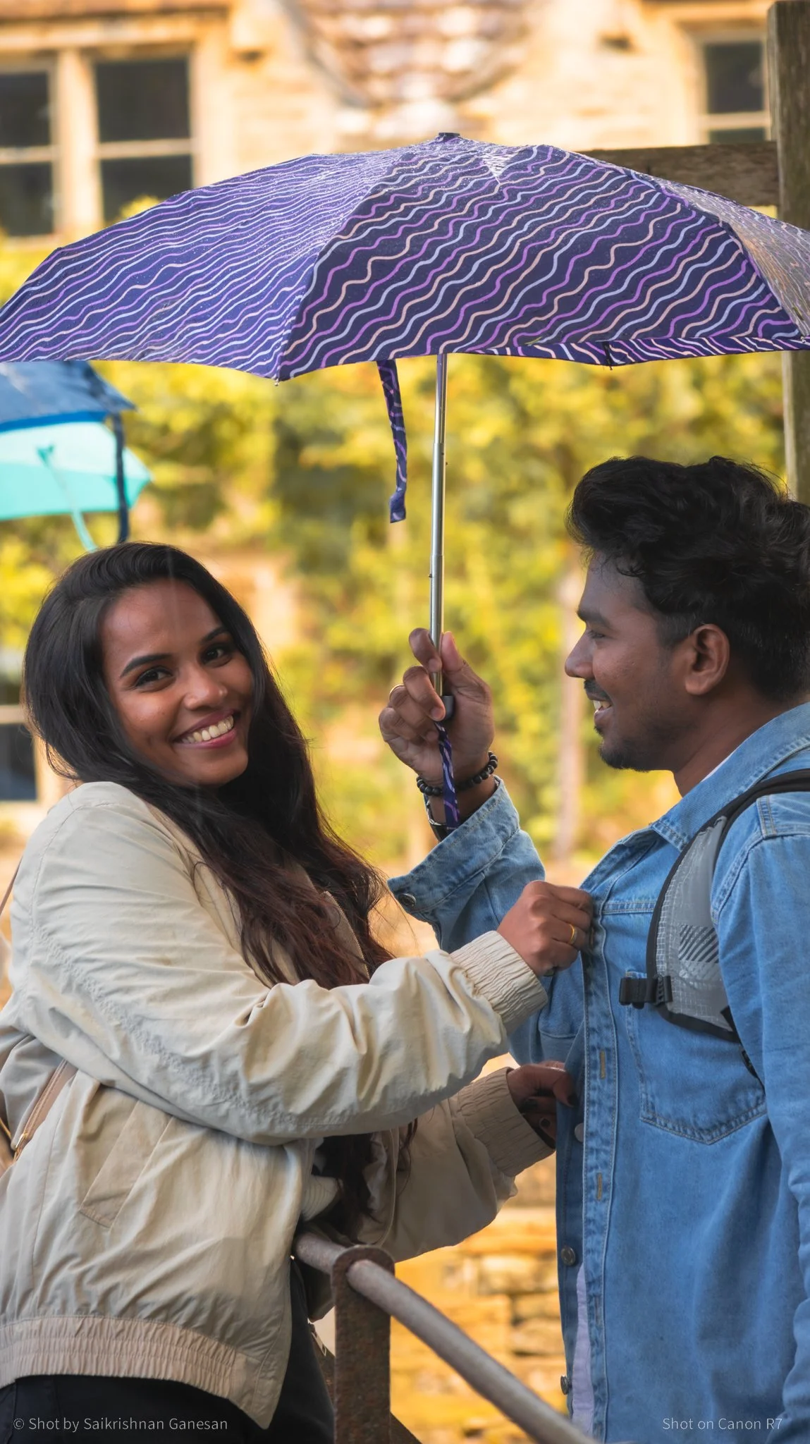 A smiling woman and a man sharing an umbrella outdoors, with autumn trees and a building in the background.