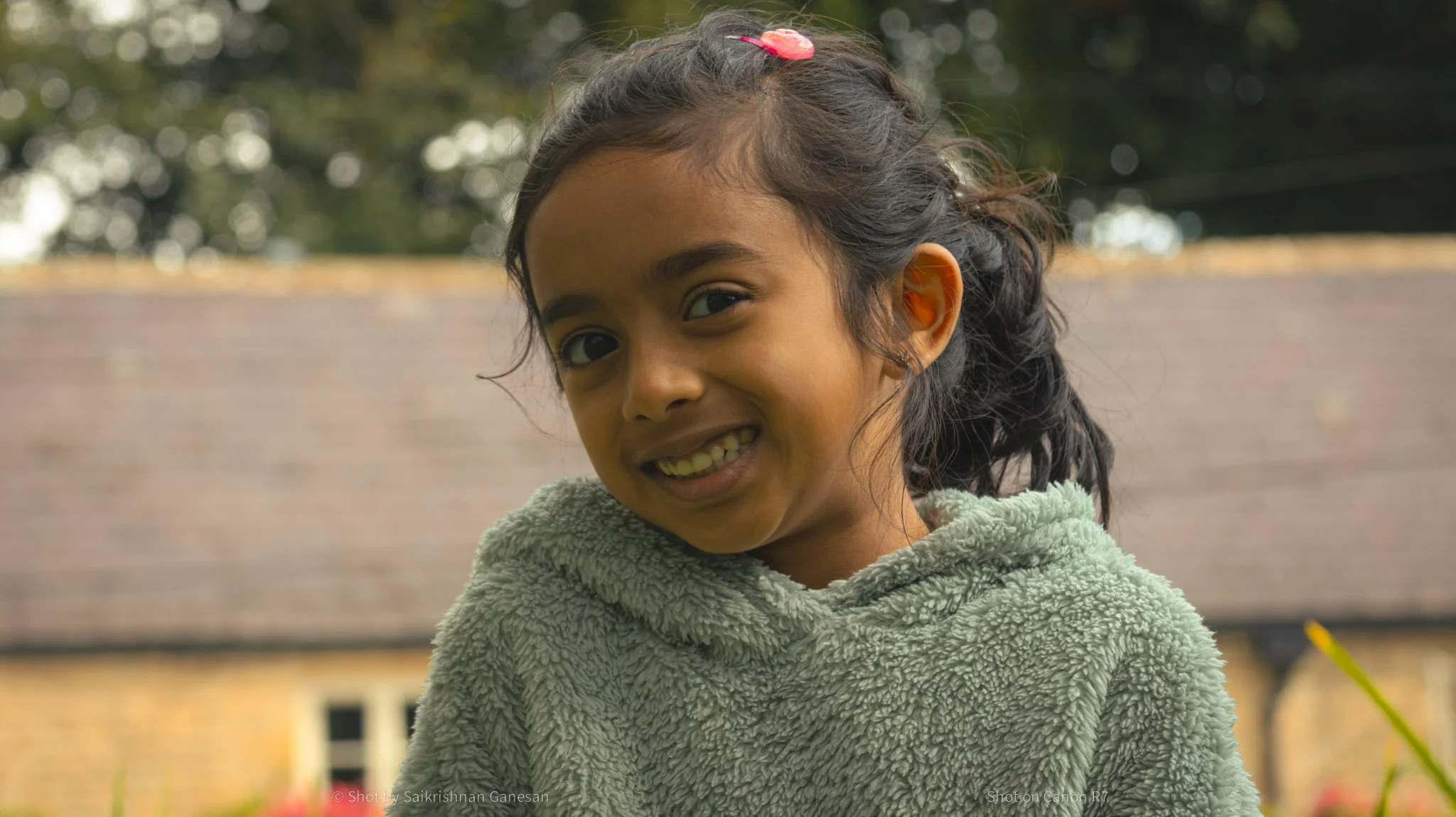 A young girl with dark curly hair, a pink hair clip, and a grey fuzzy hooded jacket, smiling outdoors with a background of trees and a brick building.