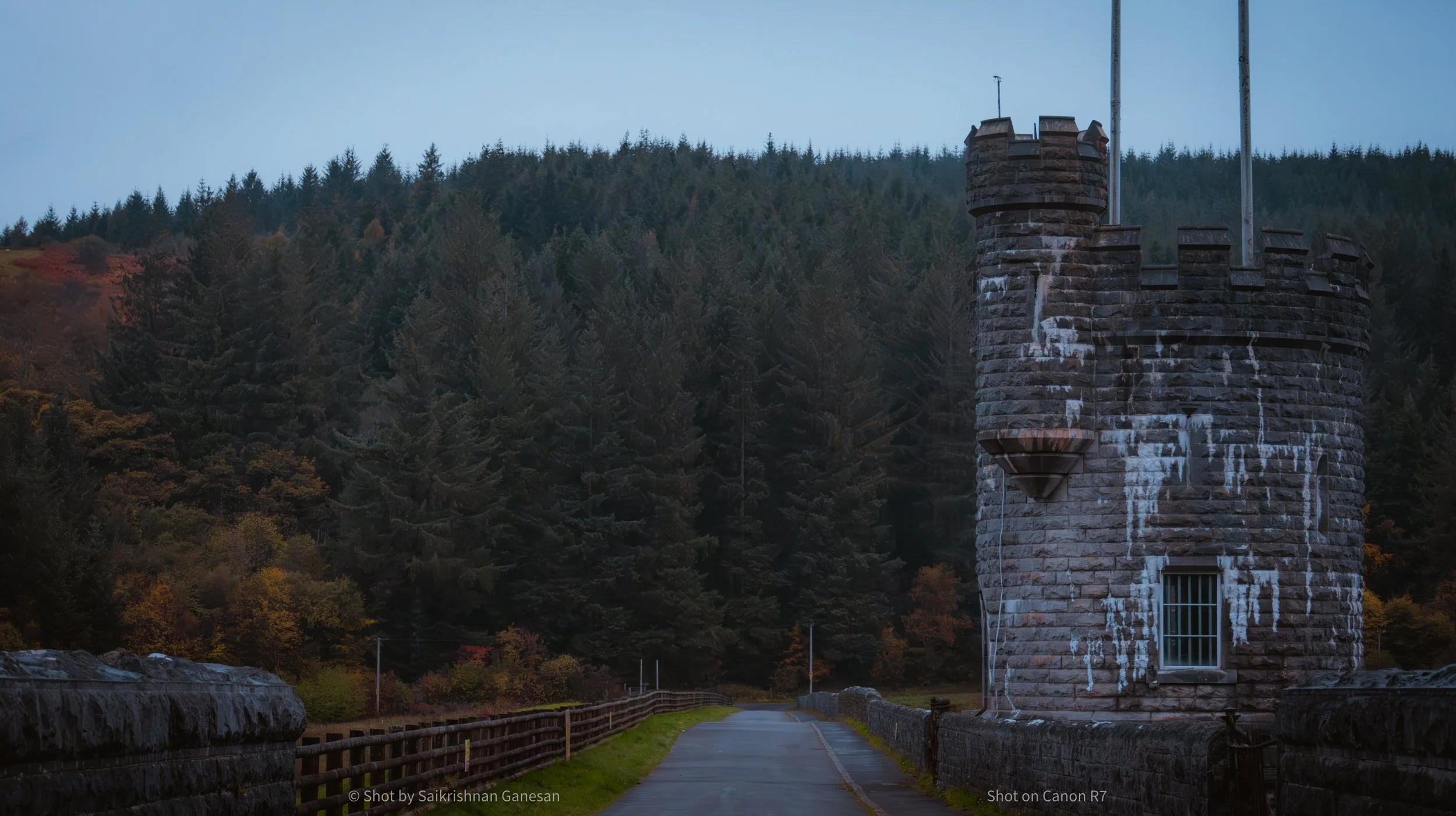 A stone castle turret on the right side of a road, with a forested hill in the background and a cloudy sky overhead.