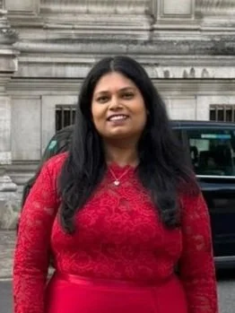 Woman with long dark hair wearing a red lace dress standing outdoors in front of a building and a parked car.